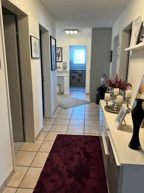 Clean, well-lit hallway with tiled flooring, white walls, and decorative items on a console table. A small rug is placed on the floor, and a bathroom is visible at the end of the hallway.