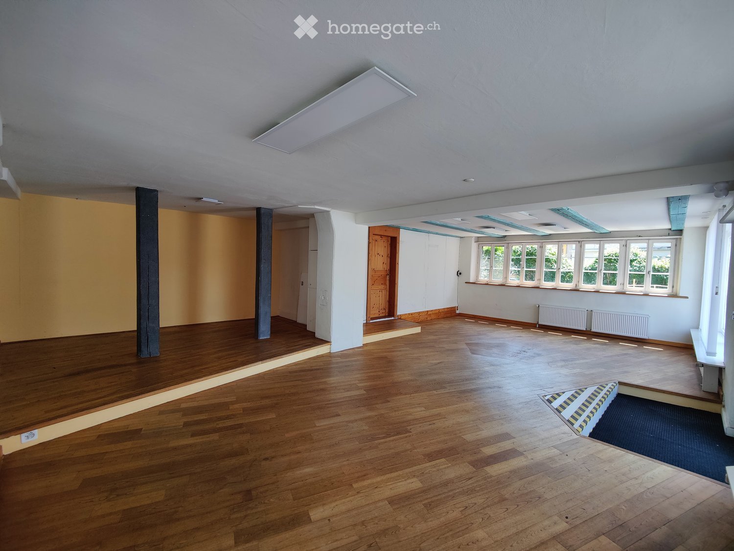 Empty room with wooden flooring, white walls, large windows, and a wooden door leading to another area.