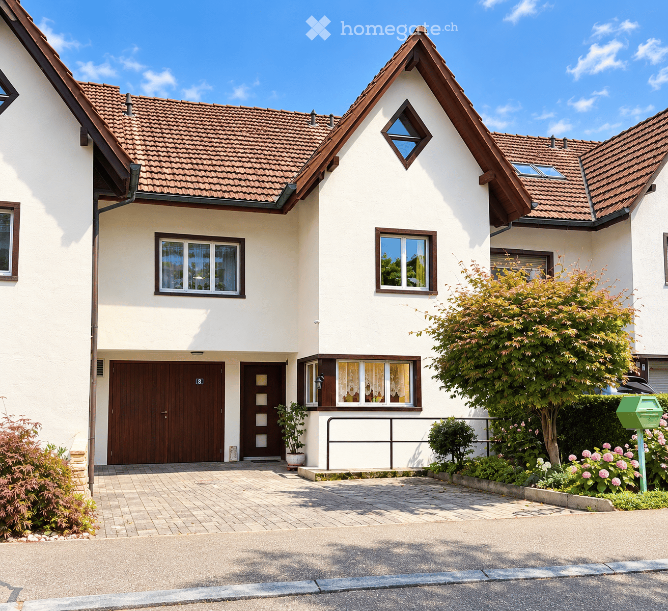 Two-story house with white walls, brown tiled roof, multiple windows, garage, and well-maintained garden with flowering plants and shrubs.