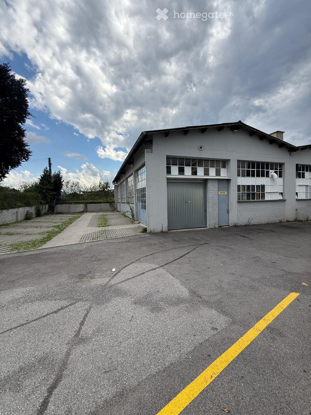 A single-story industrial building with a large garage door, multiple windows, and a paved parking area.