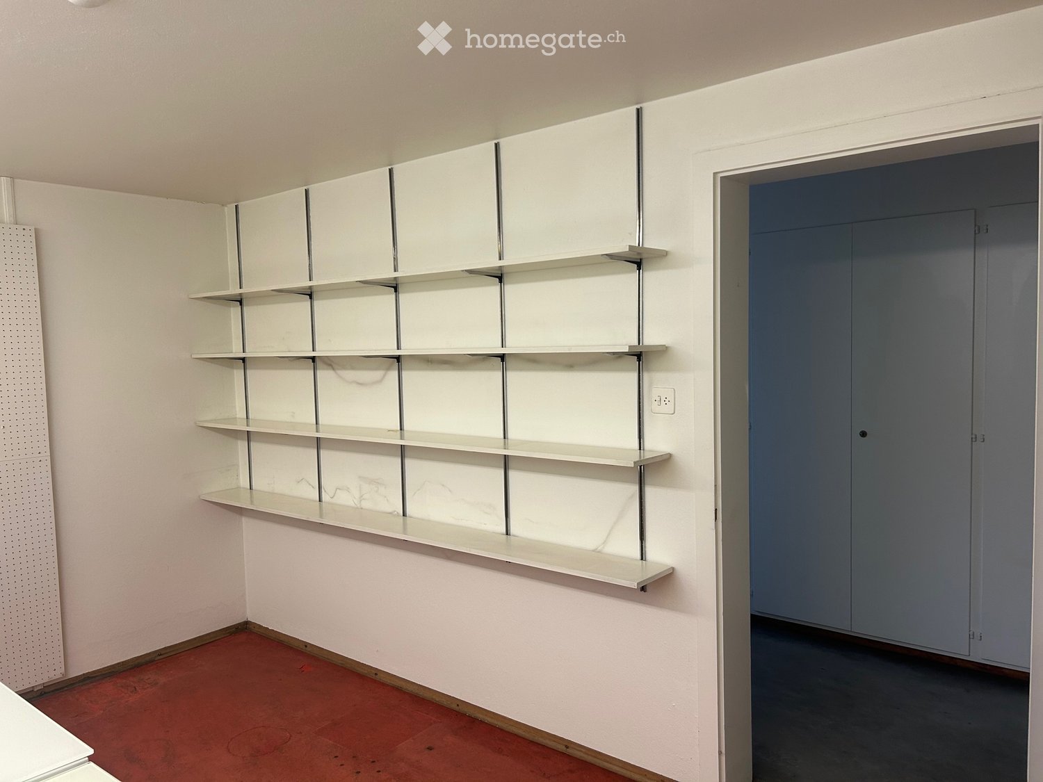 Empty room with white wall-mounted shelves, red carpeted floor, and a closed grey door leading to another space.
