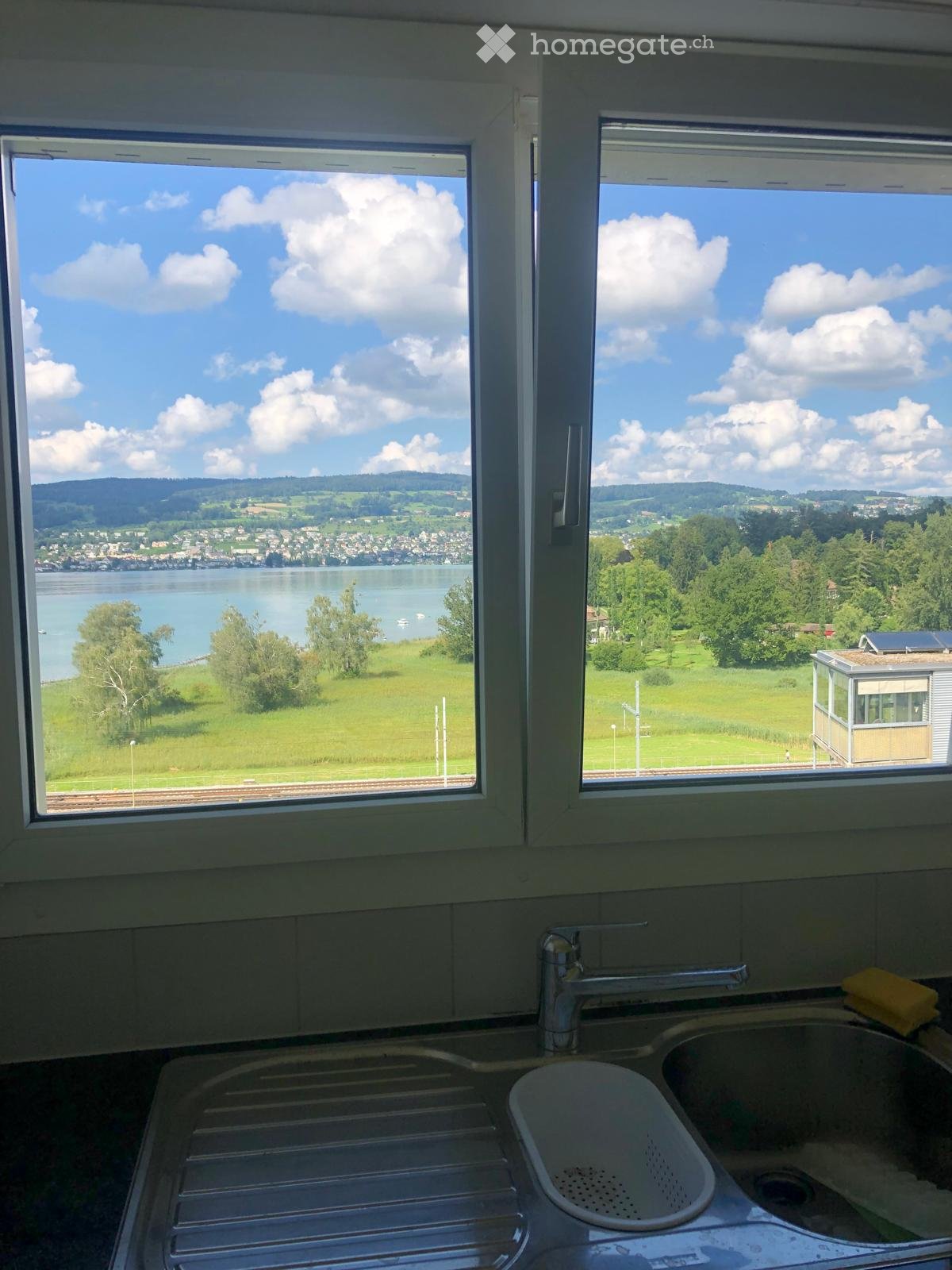 Kitchen with a double sink, faucet, and a dish rack, with large windows offering a scenic view of a lake and surrounding landscape.