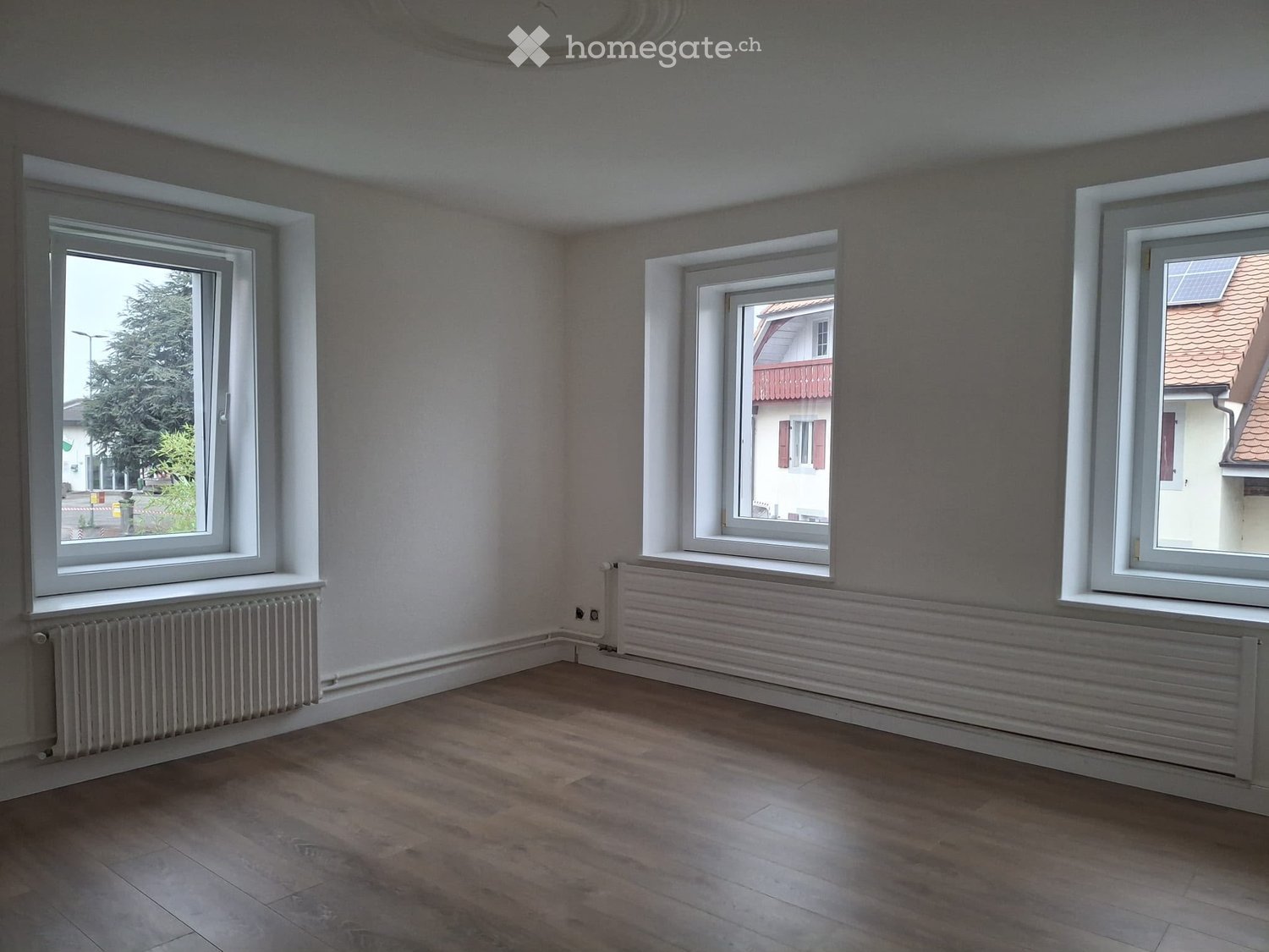 Empty room with wooden flooring, light grey walls, three windows, and radiators beneath each window.