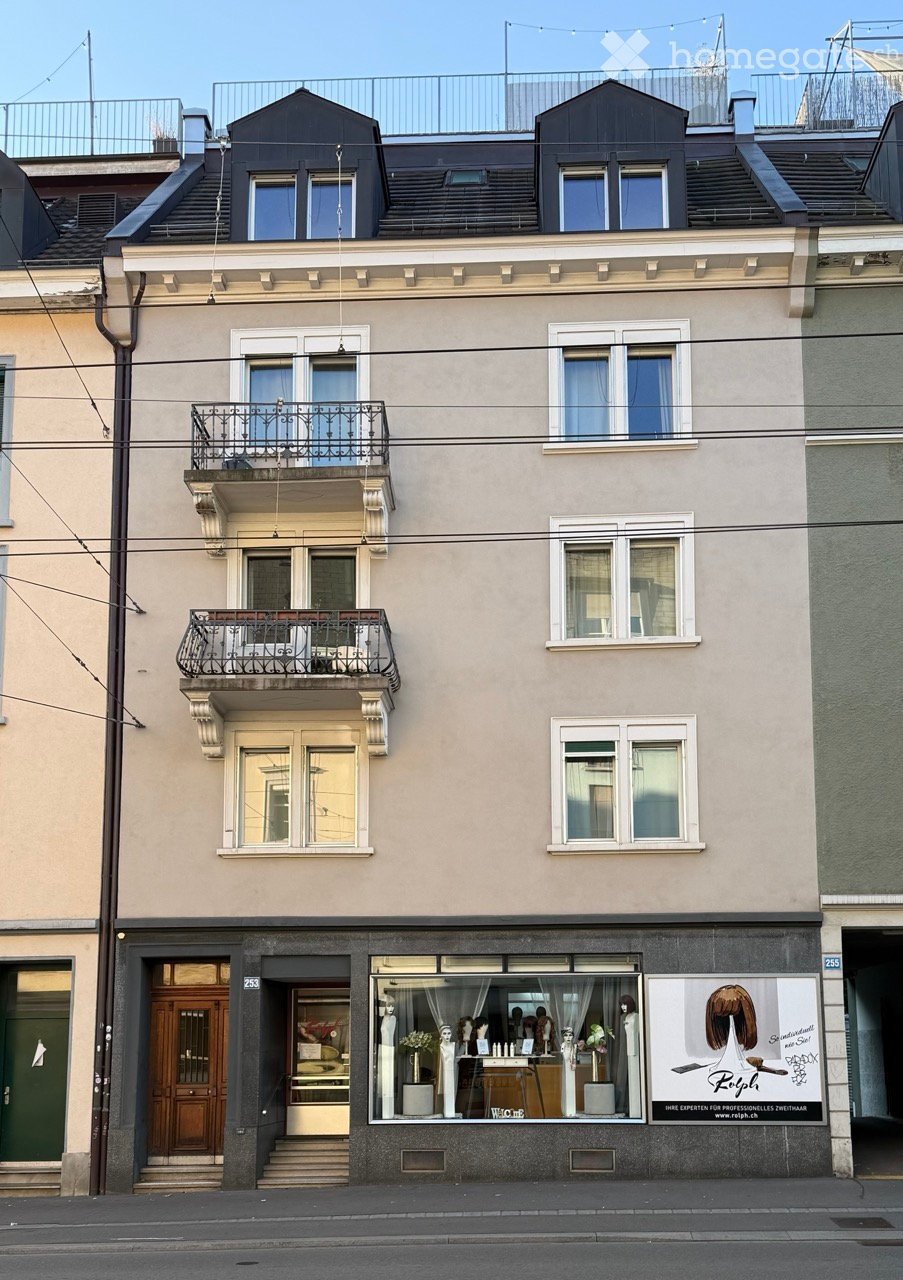 Four-story residential building with beige facade, multiple windows, two balconies, and a storefront on the ground floor.