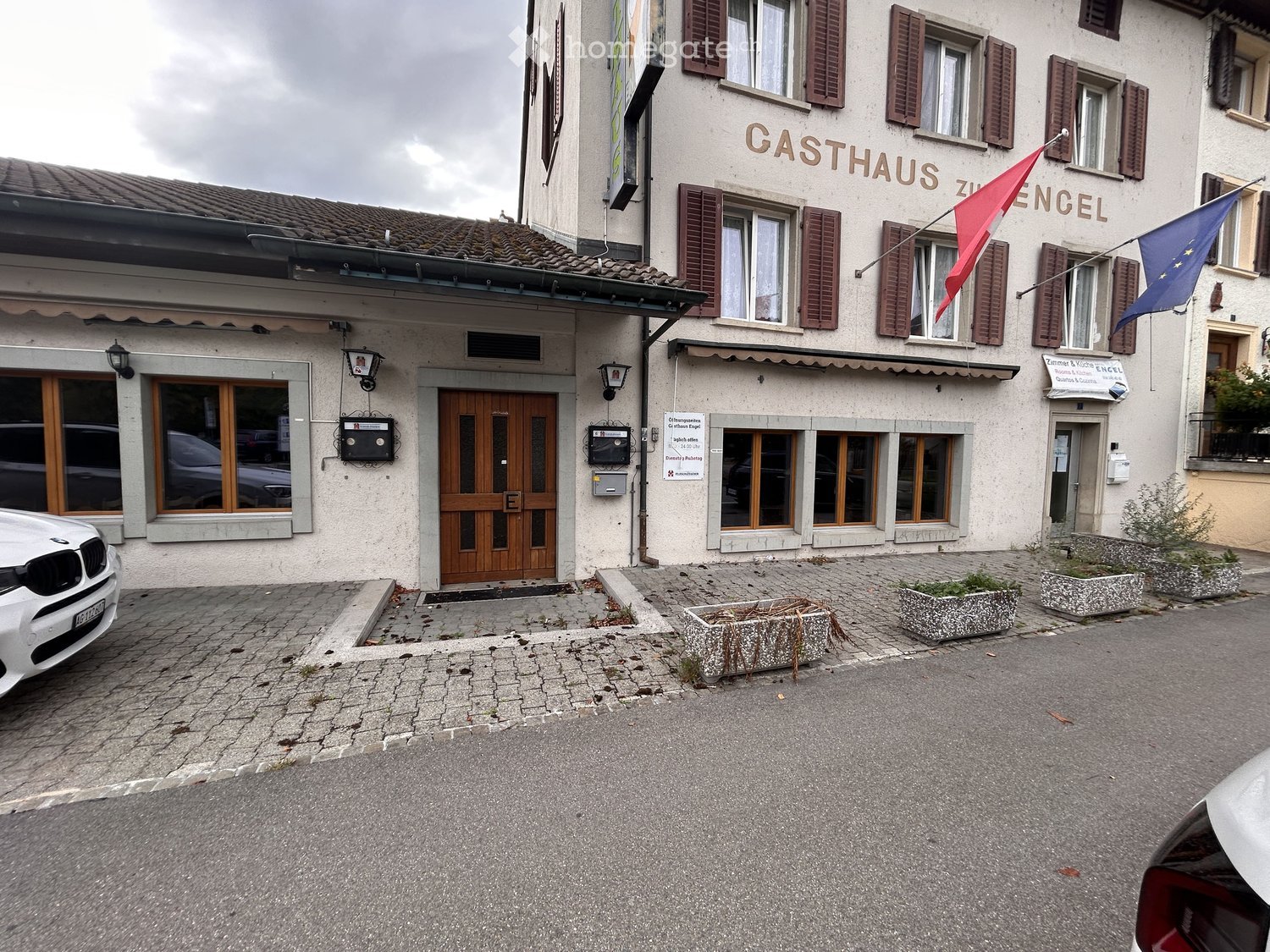 Two-story building with a sign reading "CASTHAUS ZUR ENCEL" and a flagpole with flags, including a red flag and a blue flag with a circle of stars. The building has a wooden door, windows with shutters, and a small paved area in front with planters.