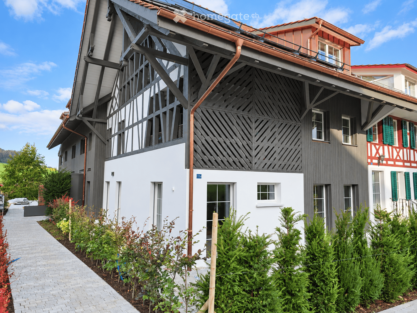 Modern two-story house with grey and white exterior, featuring a distinctive wooden lattice facade on the upper level and a red balcony section on the right. The house has multiple windows, a solar panel on the roof, and is surrounded by well-maintained g
