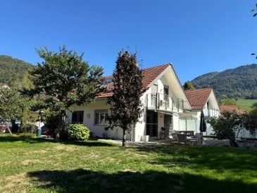 Two-story house with a red-tiled roof, white exterior, large windows, and a spacious lawn with trees and outdoor seating.