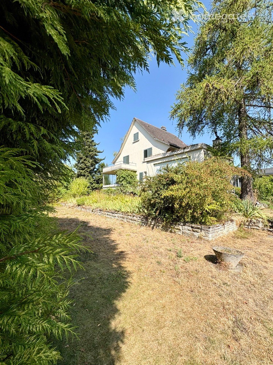 A well-maintained garden area with a gravel pathway leading to a white house with a sloped roof and large windows. The garden is surrounded by tall evergreen trees and shrubs, creating a serene and private setting.