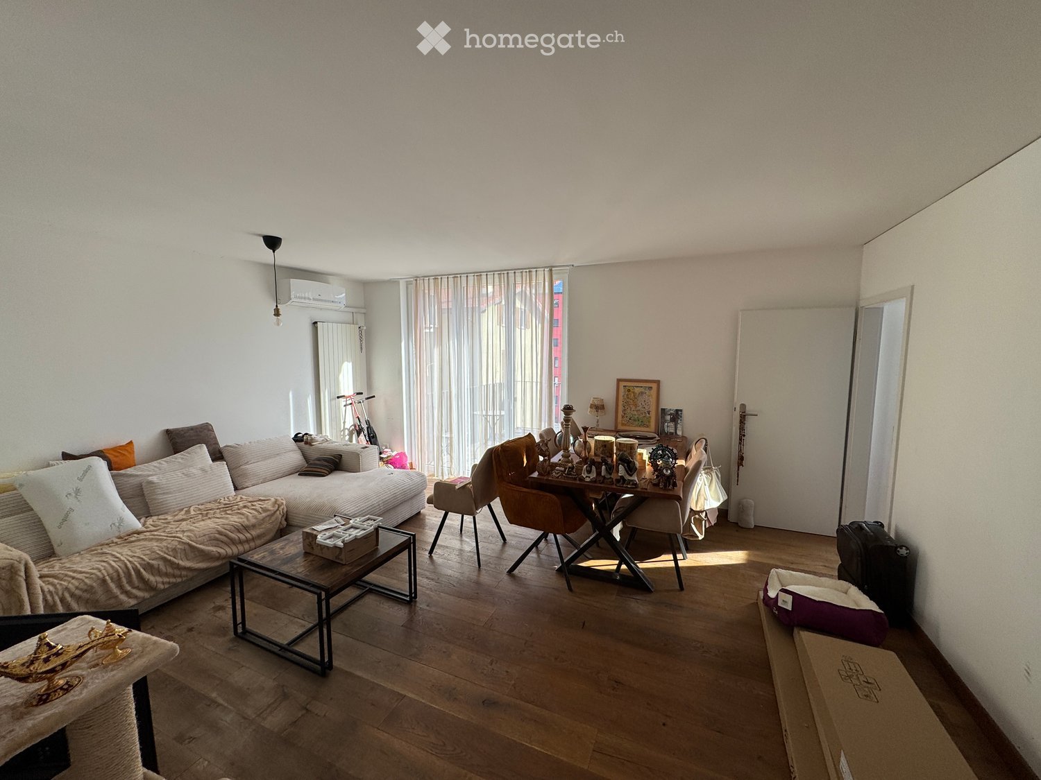 A modern living room with a sofa, coffee table, dining table, chairs, and a cat bed. The room features wooden flooring, large windows with sheer curtains, and a view of the city.