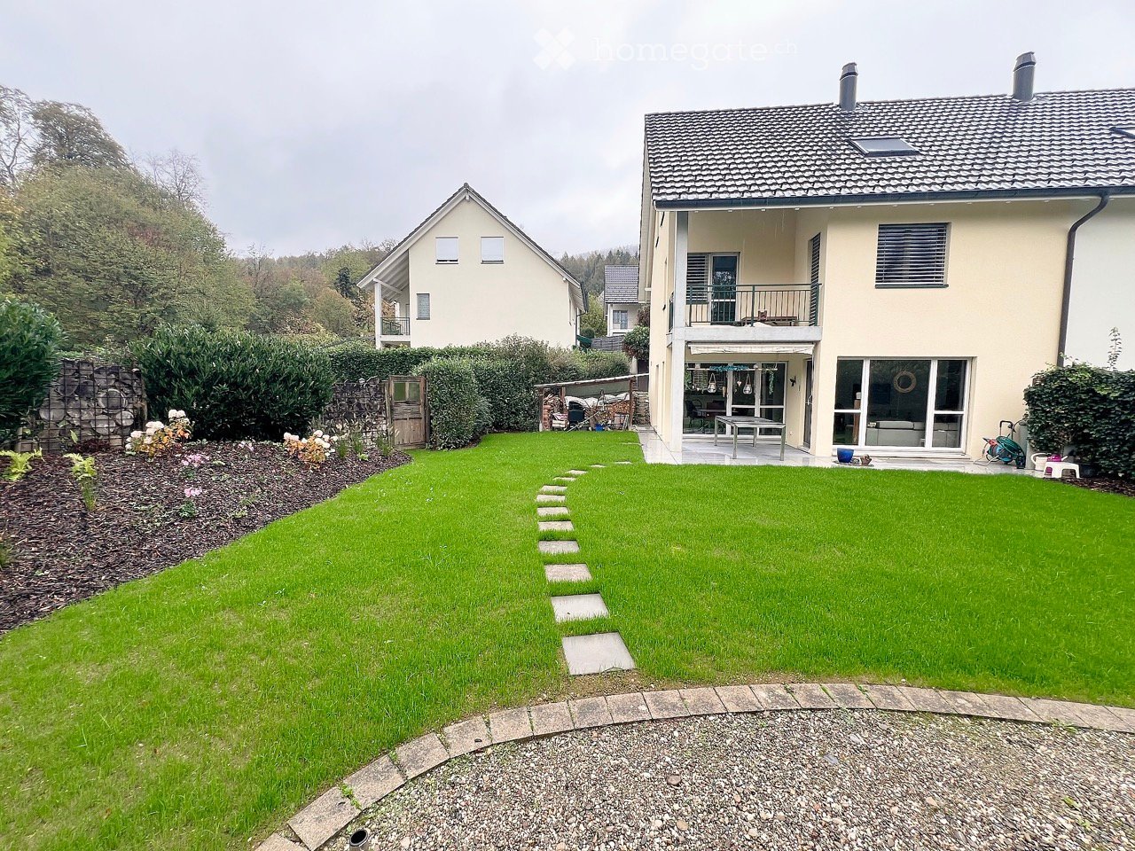 Well-maintained garden with a stone pathway leading to a modern house, featuring a patio area with outdoor furniture and a view of neighboring houses.