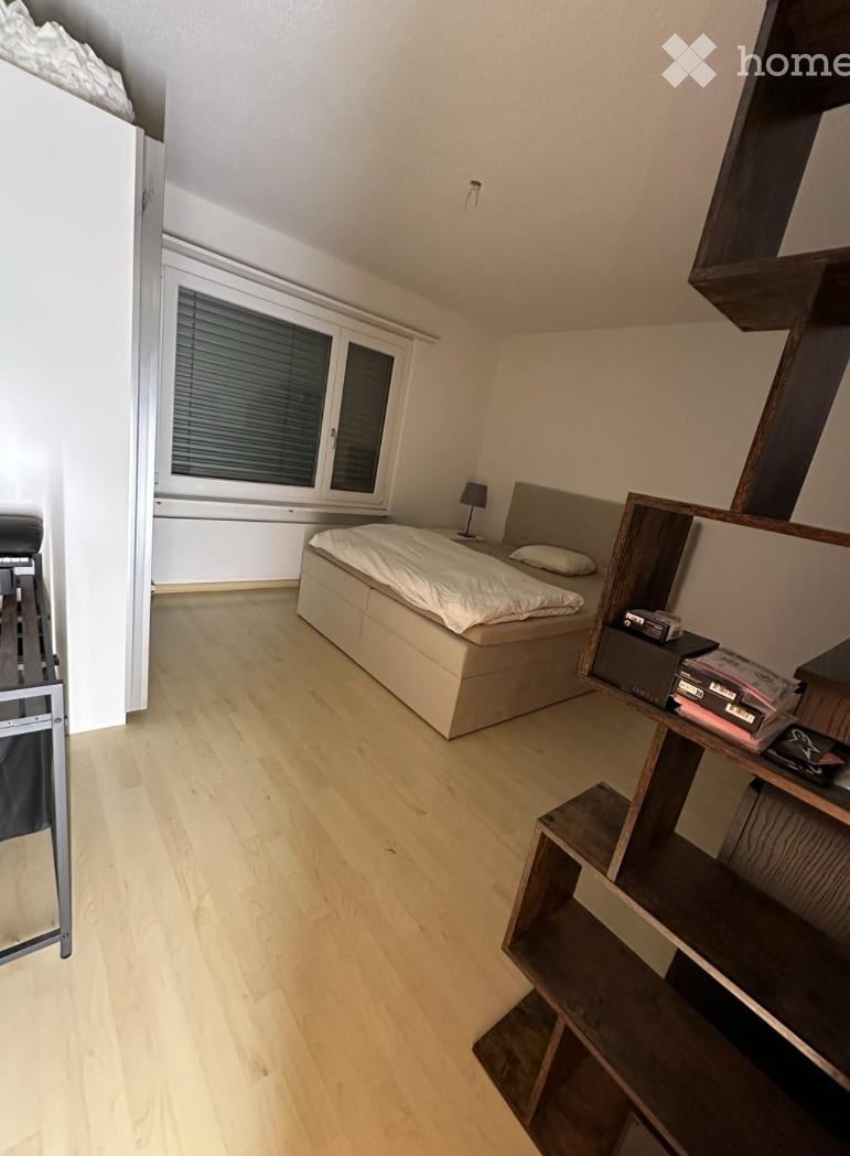 A modern, minimalist bedroom with light wooden flooring, a large window with blinds, a neatly made bed, a nightstand with a lamp, and a wooden shelf with books and decorative items.