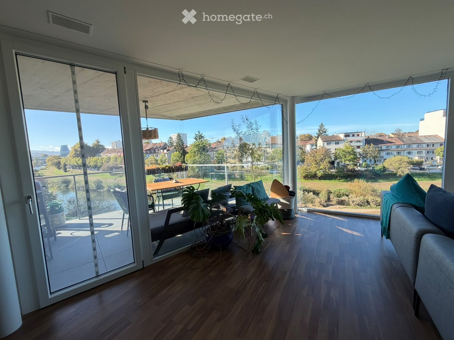 Bright living room with wooden flooring, large glass doors leading to a balcony, sofa, table, chairs, and potted plants.