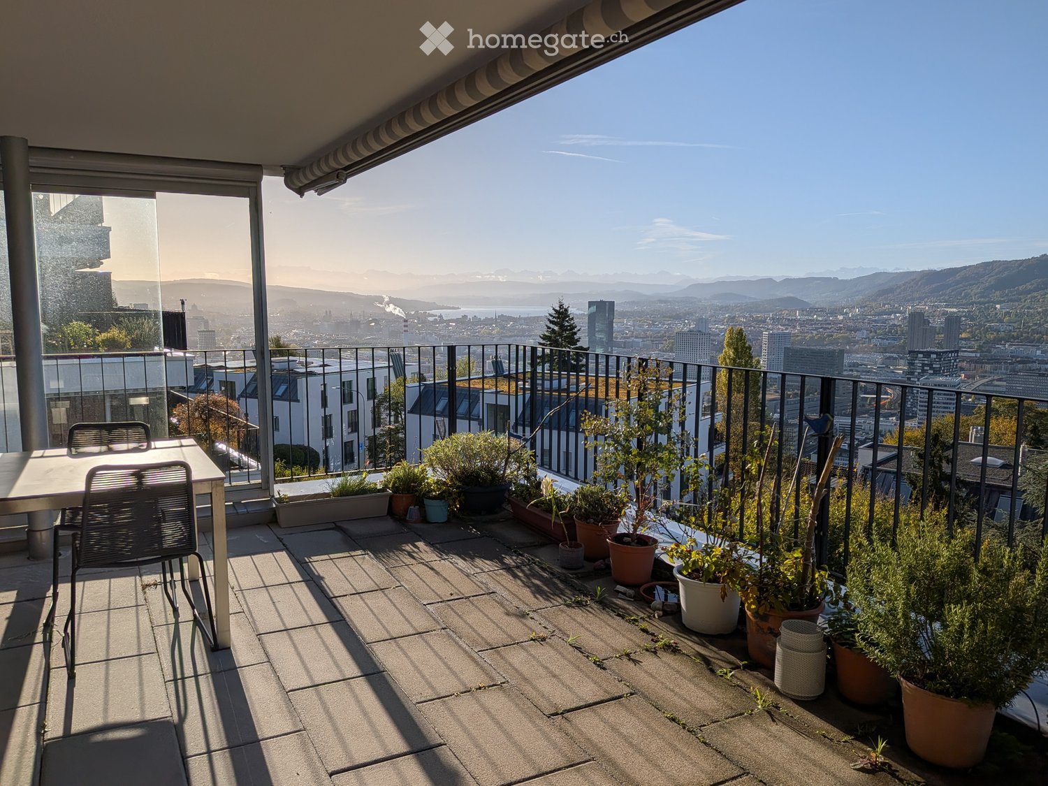 Balcony with a cityscape view, featuring a table, chairs, and several potted plants arranged along the railing.