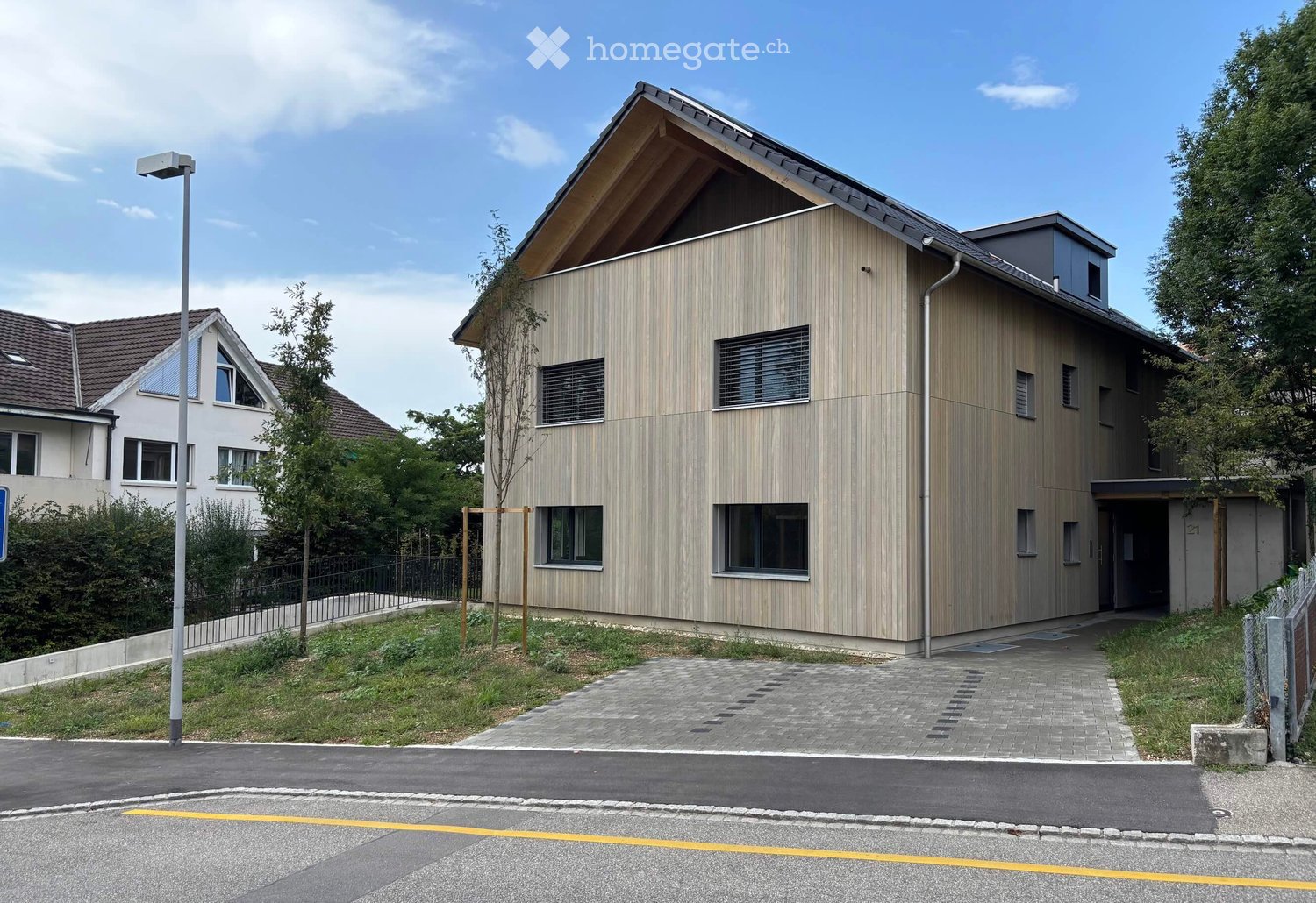 Modern two-story house with a wooden facade, large windows, and a sloped roof. The house features a small balcony and is surrounded by greenery and a paved driveway.