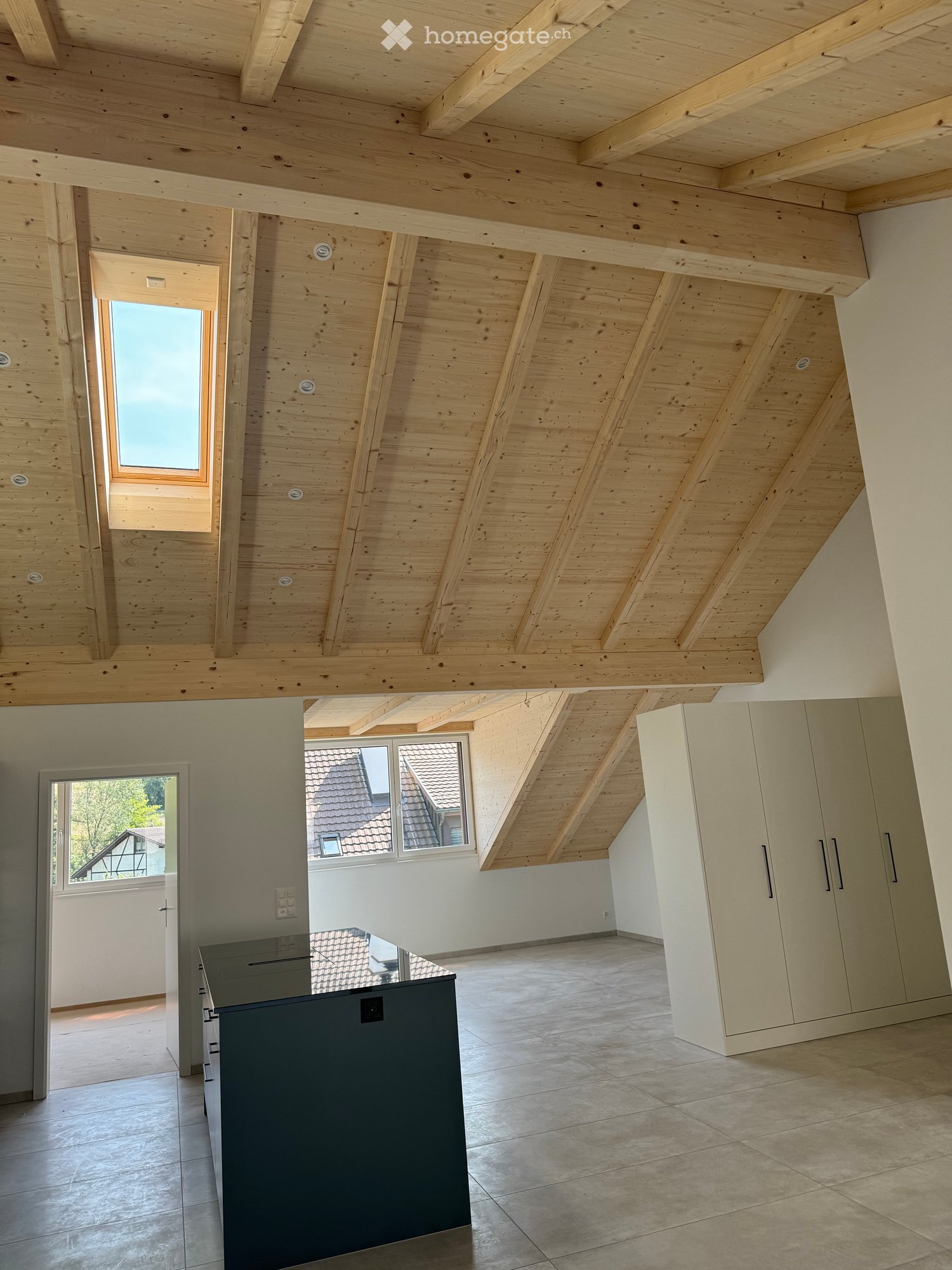 An empty attic space with exposed wooden beams and slanted ceilings, featuring large windows and built-in storage cabinets.