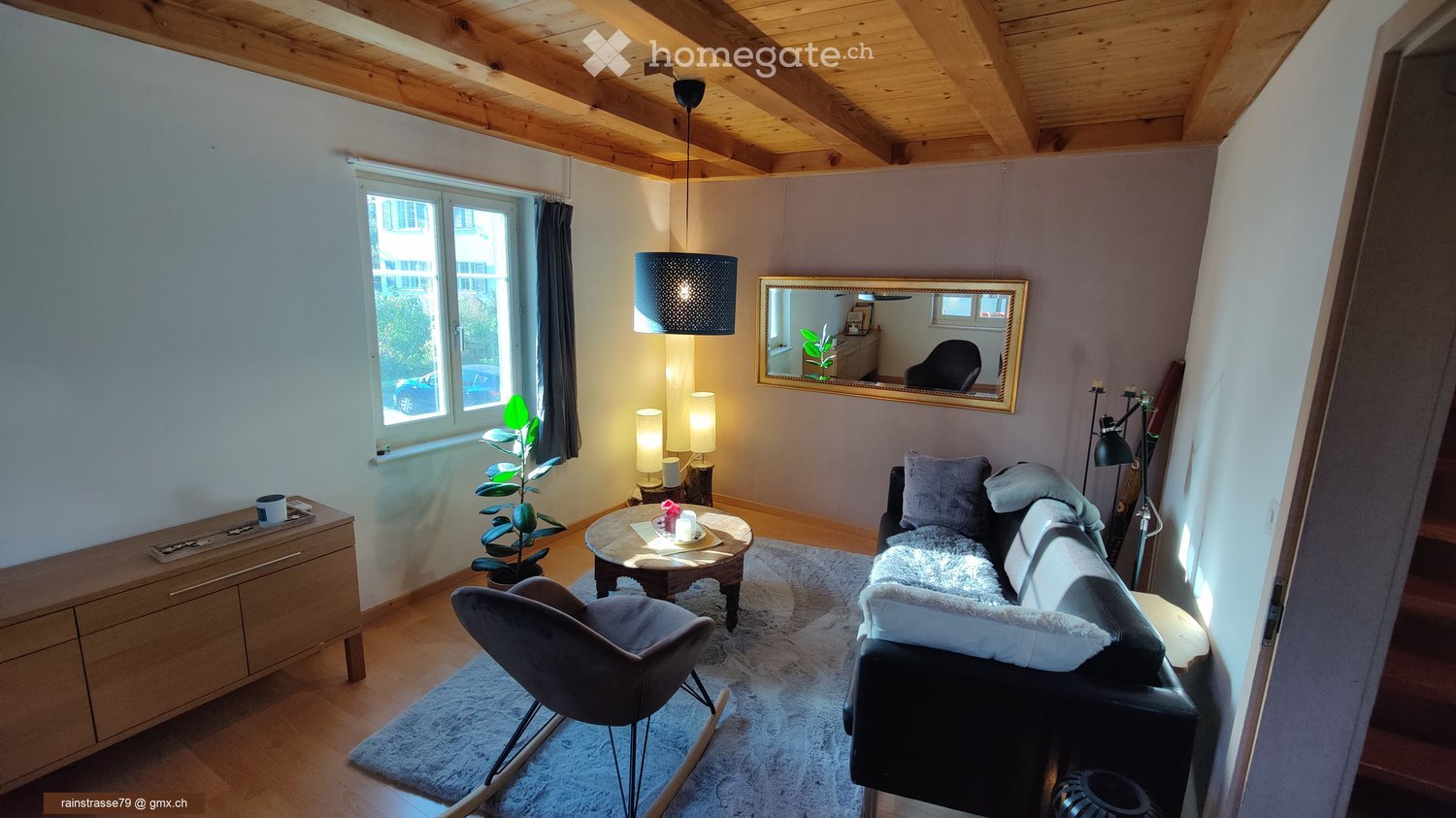 Cozy living room with wooden ceiling beams, large window, grey sofa, coffee table, armchair, and decorative items.