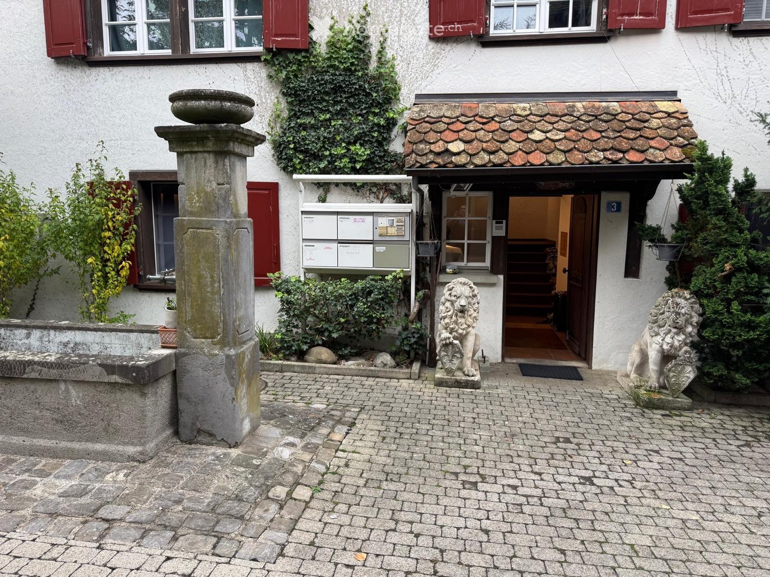 Entrance to a two-story building with a stone-paved courtyard, red shutters, ivy-covered walls, and two stone lion statues flanking the doorway.