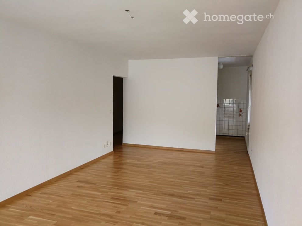 Empty room with wooden flooring and white walls, featuring a doorway and a glimpse of a tiled bathroom area.