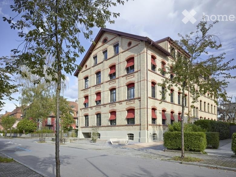A multi-story brick building with red awnings, large windows, and a symmetrical facade. The building is surrounded by trees and greenery, with a paved area in front.