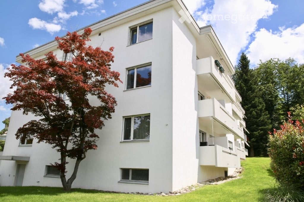 Modern white multi-story building with multiple windows and balconies, surrounded by greenery and a well-maintained lawn.
