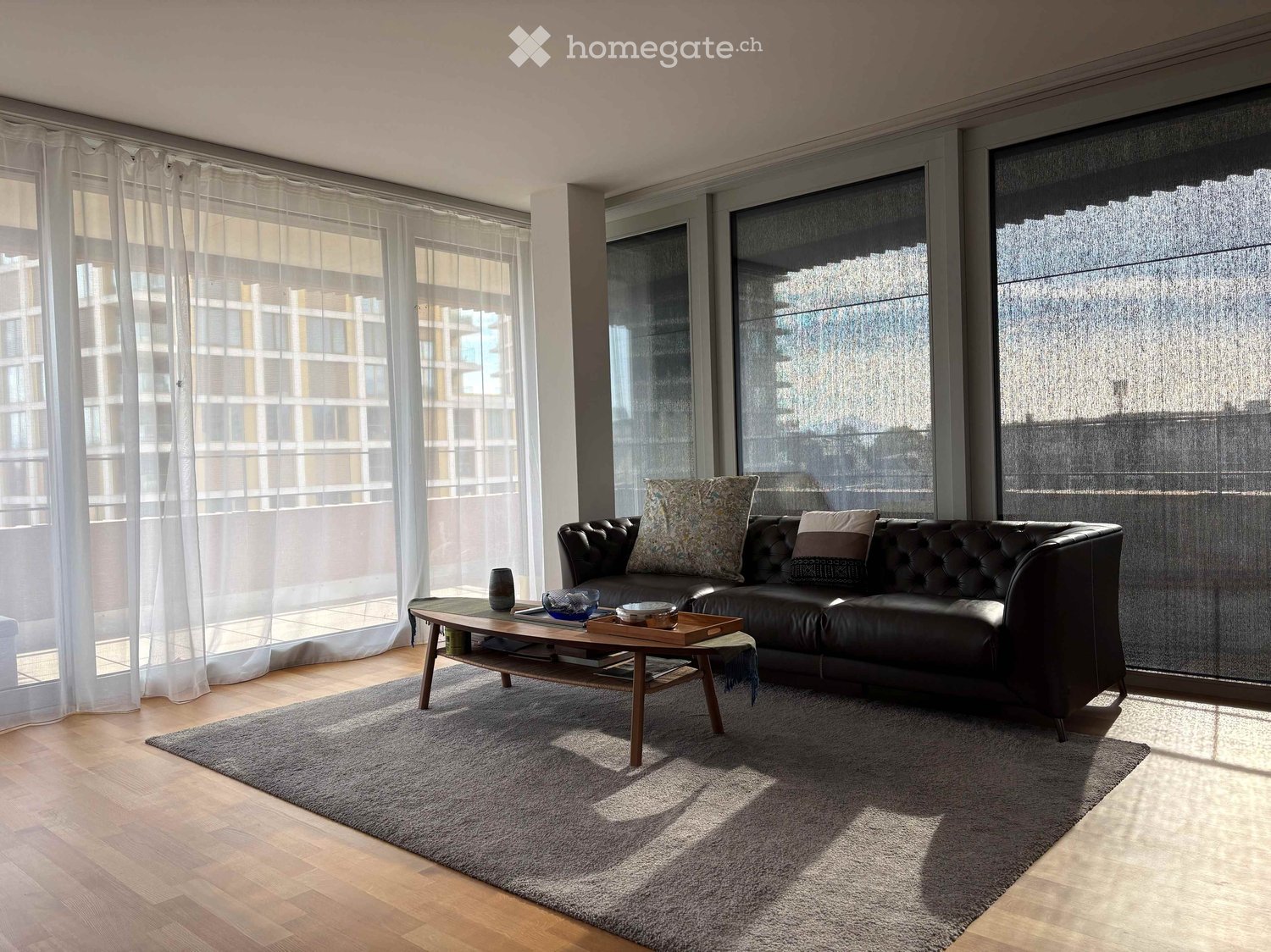 Modern living room with large windows, a black leather sofa, a wooden coffee table, and a grey rug.
