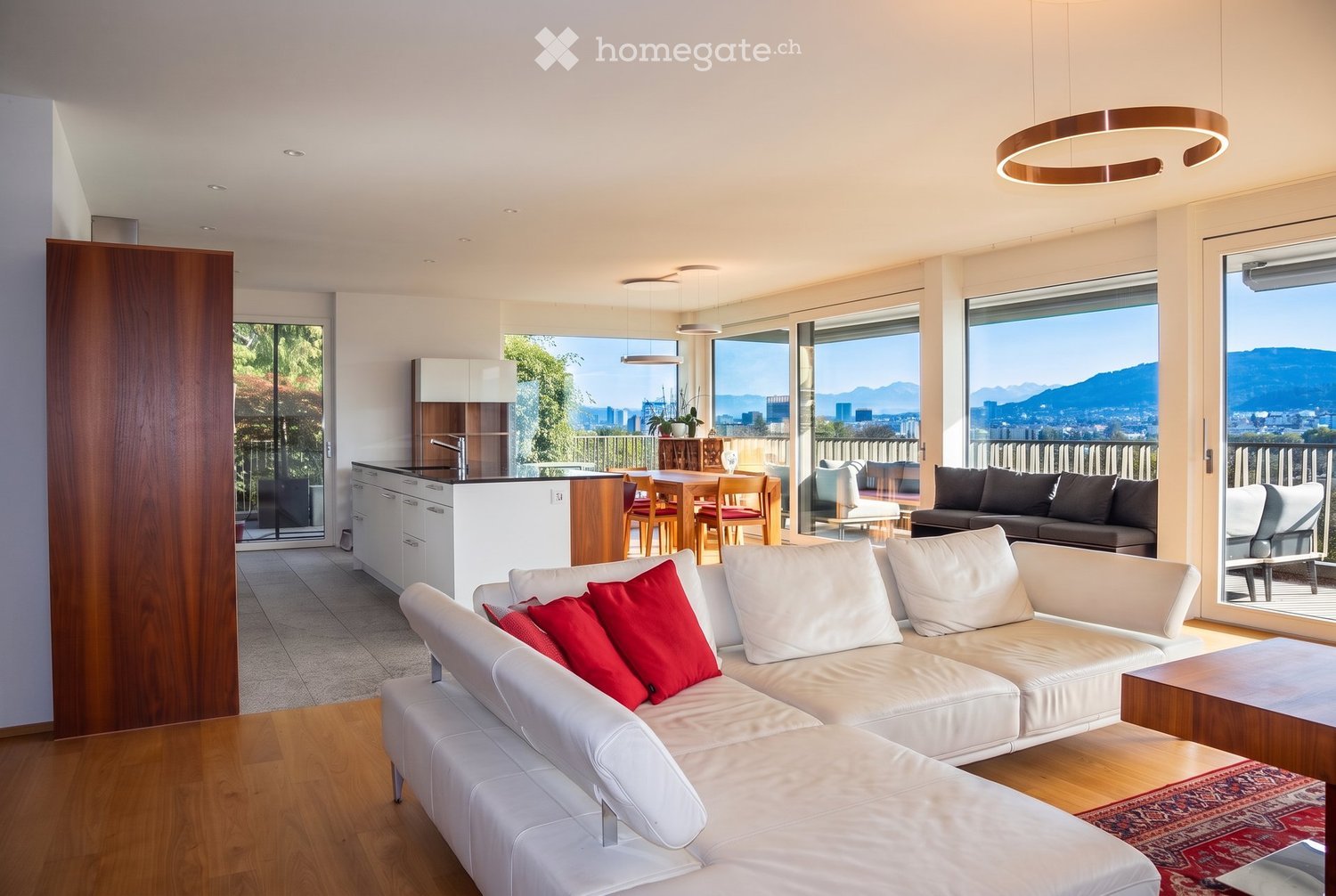 Modern living and dining area with white leather sofa, red throw pillows, coffee table, dining table with chairs, kitchen area, large windows offering a scenic view, and pendant lighting.