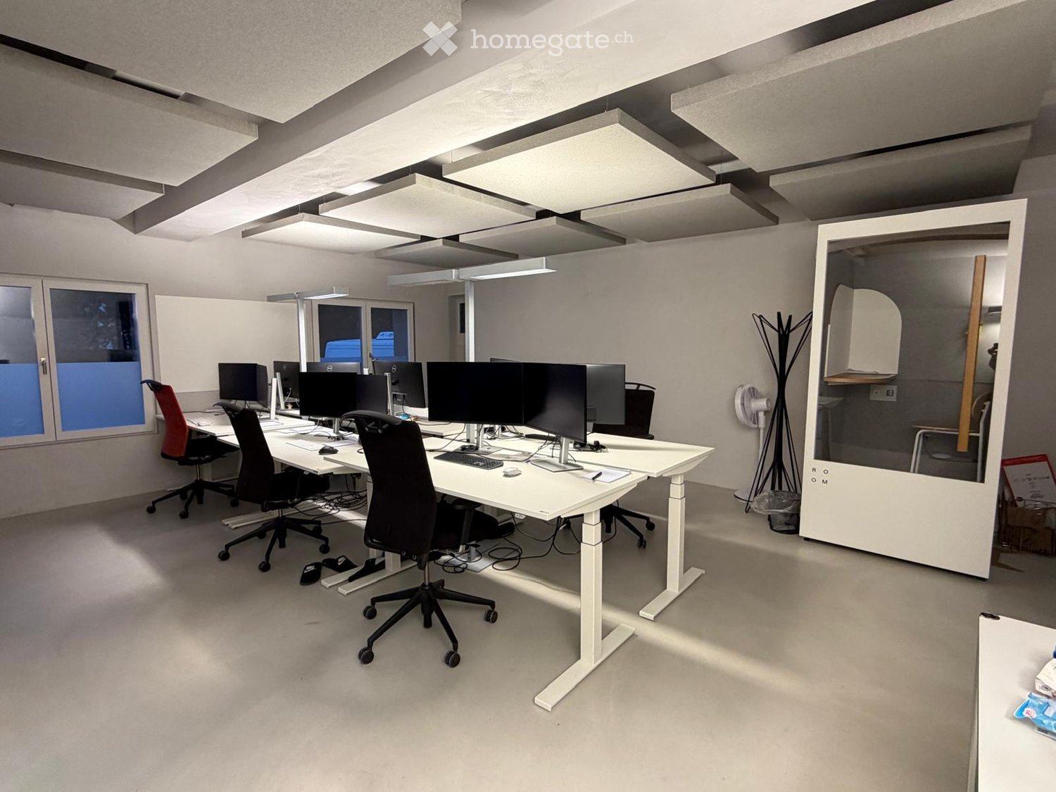Modern office space with white desks, ergonomic chairs, multiple computer monitors, keyboards, and a standing fan. Large windows provide natural light, and a screen divider separates workspaces.