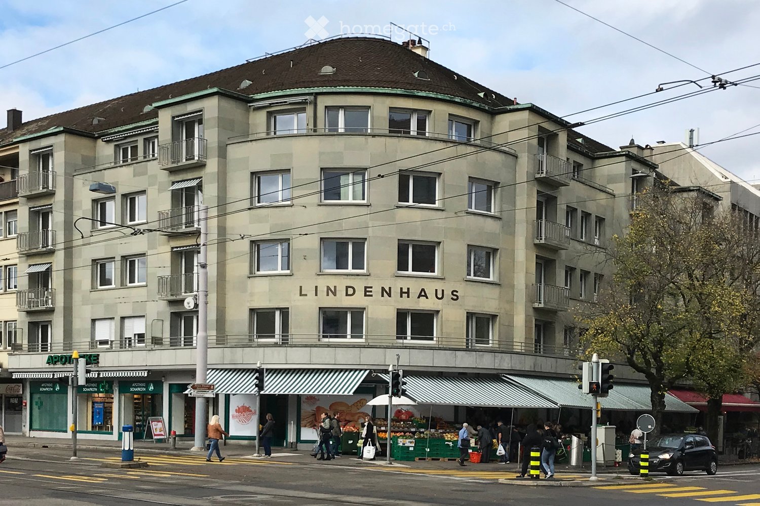 Multi-story building with a rounded roof and balconies, featuring the sign "LINDENHAUS" on the facade. The ground floor includes shops and a market area with people walking and shopping.