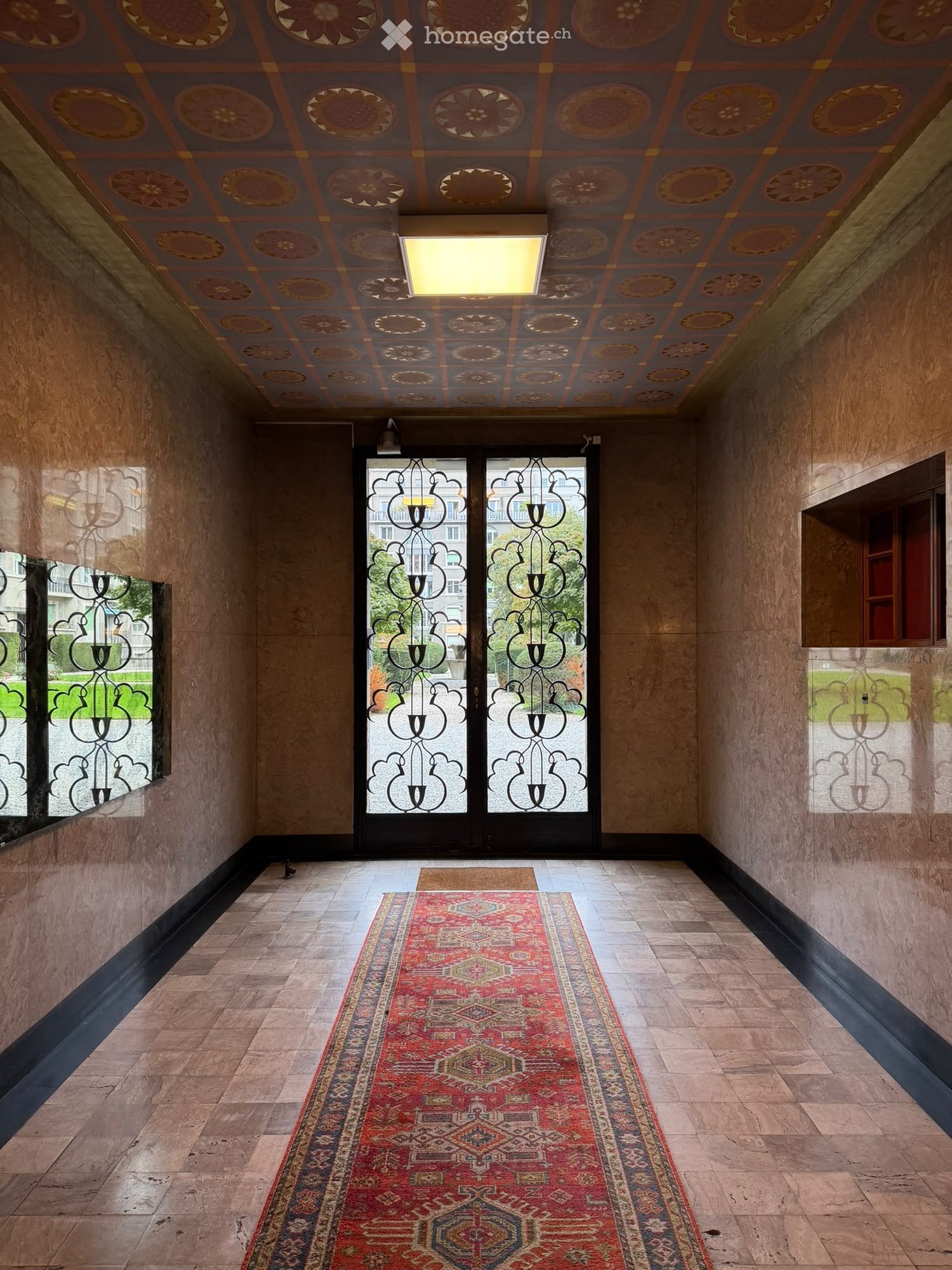 Ornate hallway with decorative ceiling, patterned rug, large double doors with glass panels, and wooden flooring.