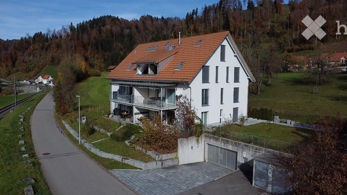 A modern two-story house with a red-tiled roof, white exterior, and a balcony on the upper floor. The house is situated on a raised ground floor with a garage and surrounded by a well-maintained garden and trees.