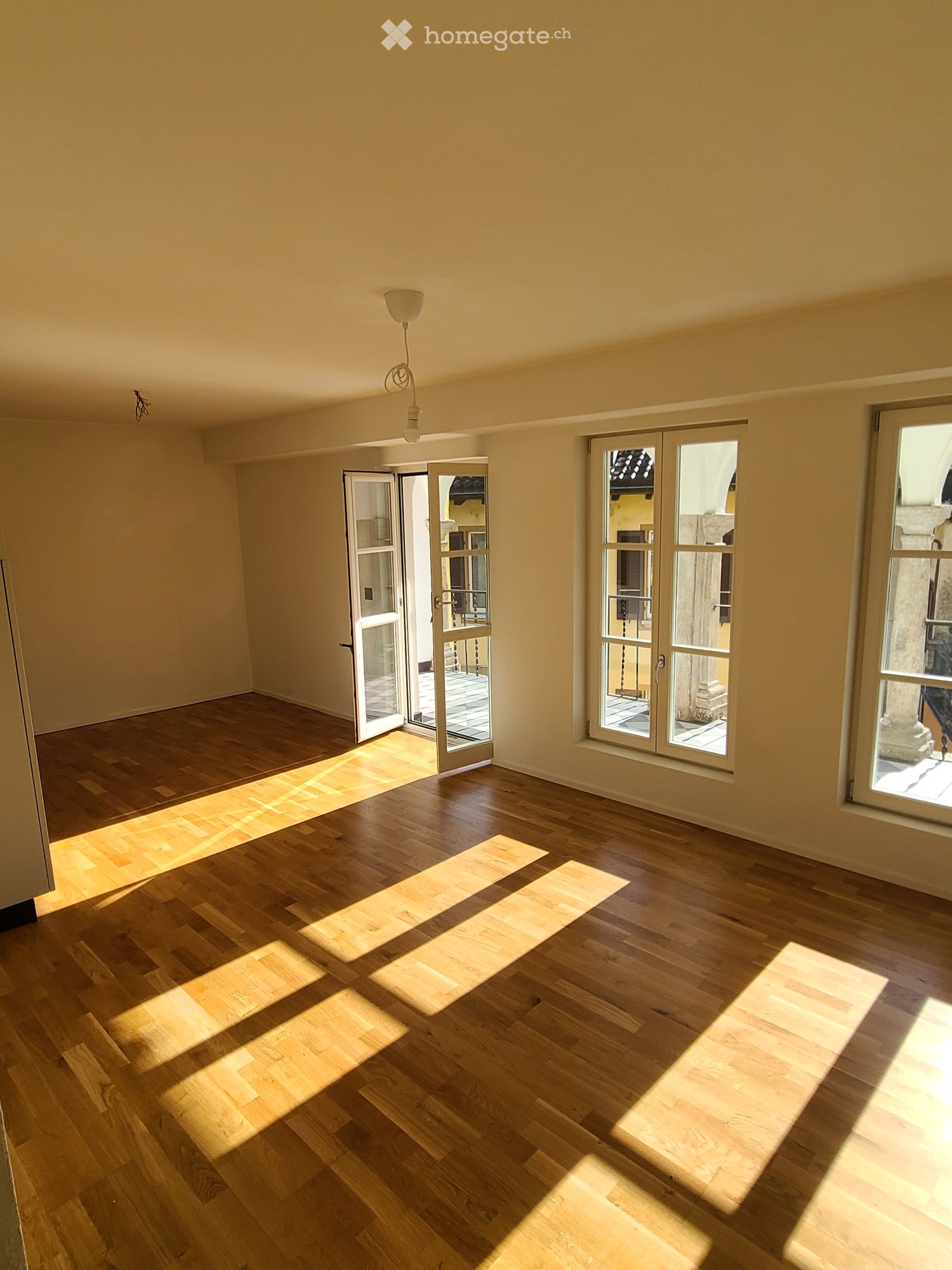 Empty room with wooden flooring, large windows, and open doors leading outside.