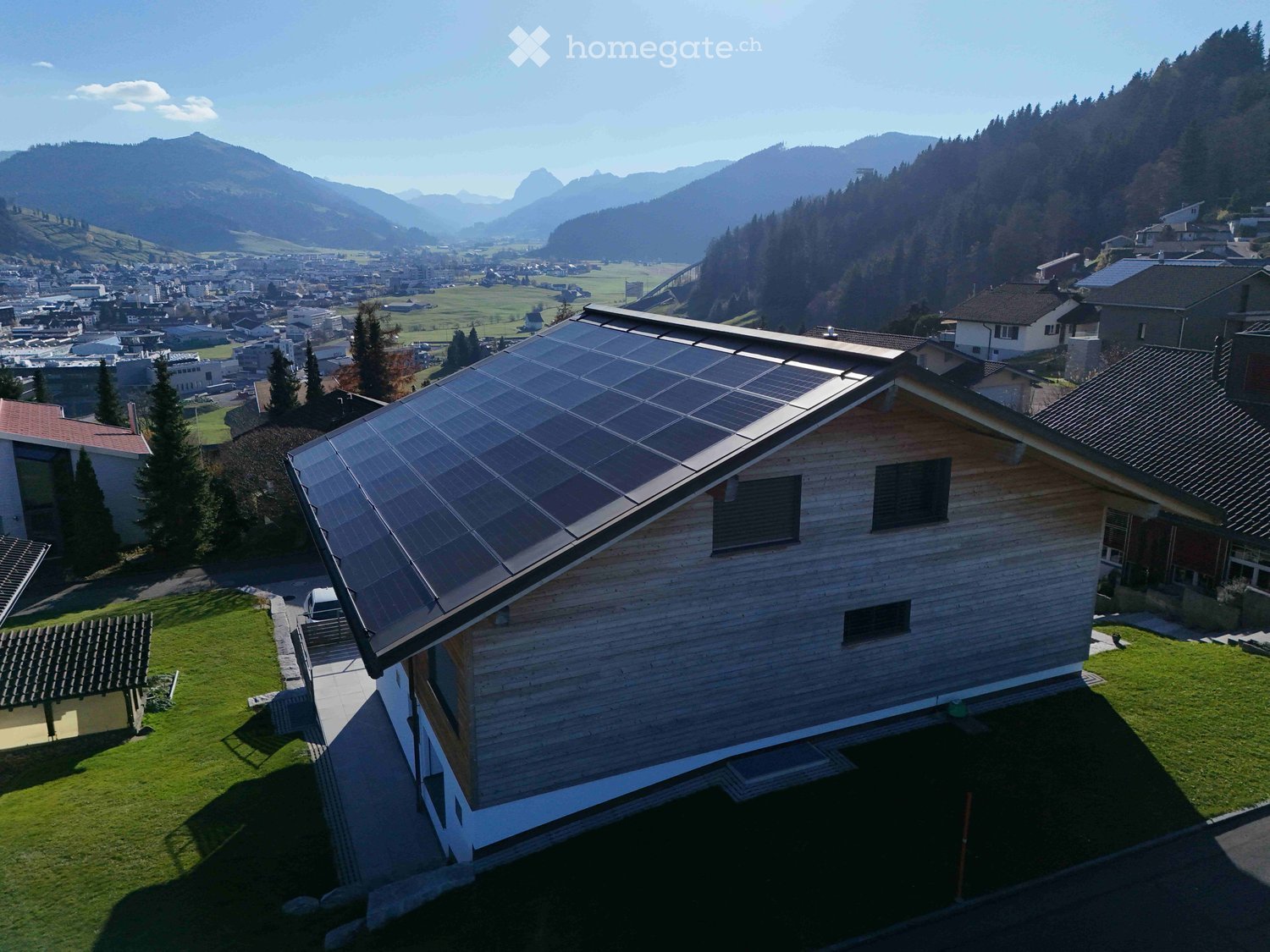 Wooden house, solar panels on the roof, located in the mountains, surrounded by a town, with a car parked in front