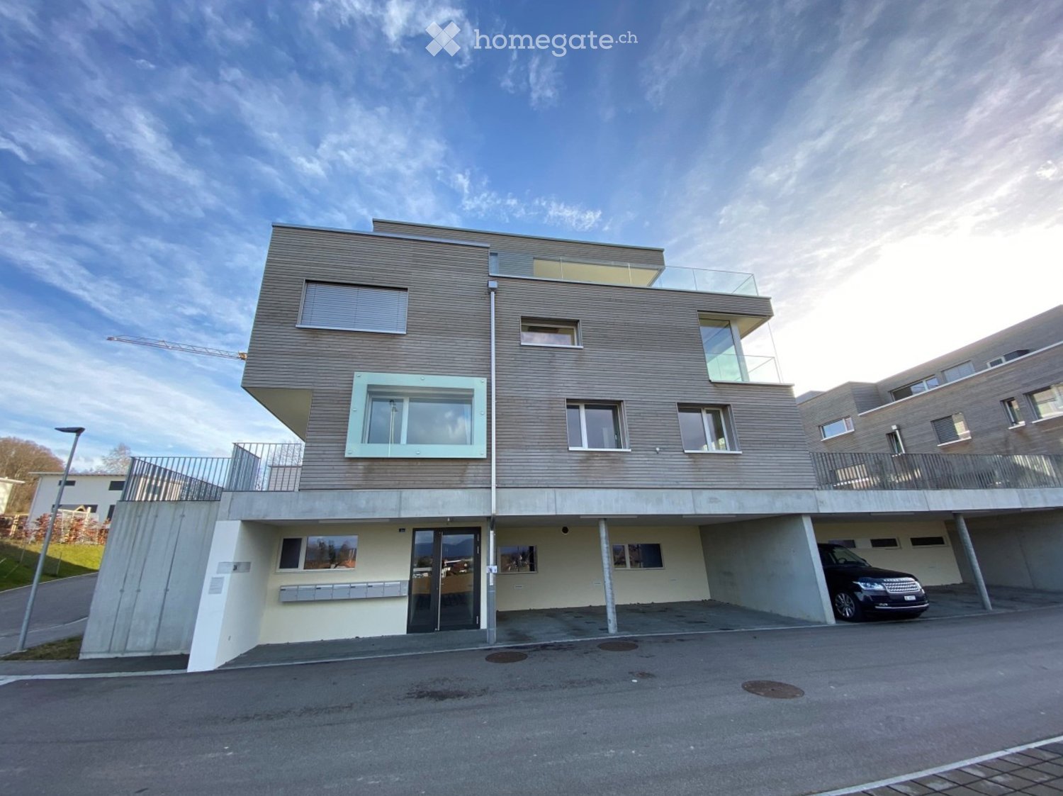 Modern two-story building with a raised ground floor, large windows, and a balcony on the upper level. A car is parked in the covered garage area.