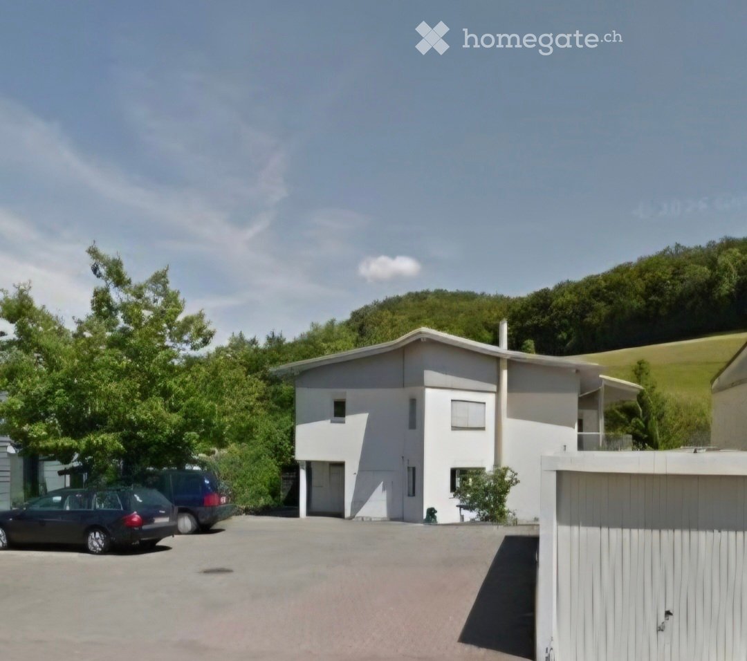 Modern two-story house with white exterior, large windows, and a flat roof, situated in a green, hilly area with a parking space in front.