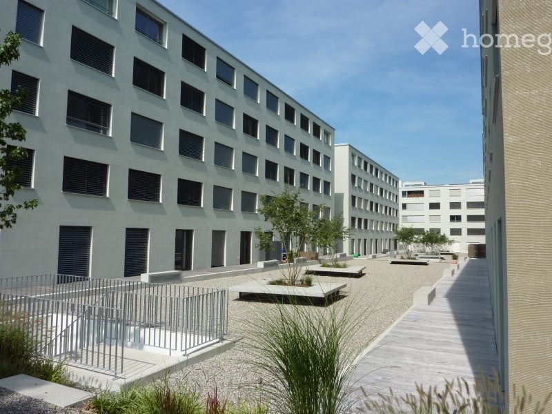 Modern multi-story building with a clean, minimalist design, featuring numerous windows and a raised ground floor with a paved courtyard and low seating areas.
