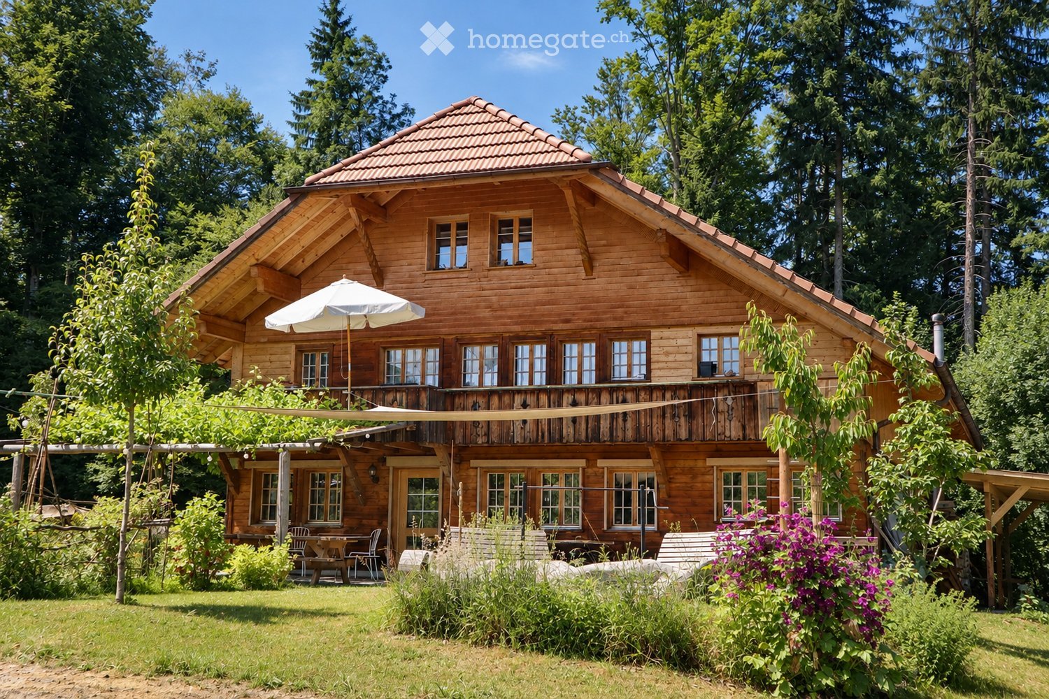 A two-story wooden house with a red-tiled roof, large windows, and a balcony. The house is surrounded by lush greenery and features outdoor seating with a table and chairs under a pergola.
