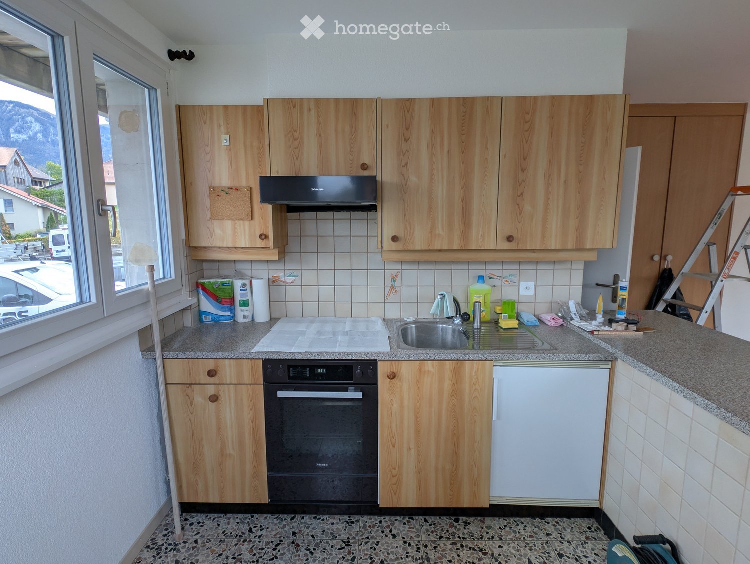 A well-lit kitchen with wooden cabinets, a stainless steel sink, and a black oven. The countertop has cleaning supplies and a broom.