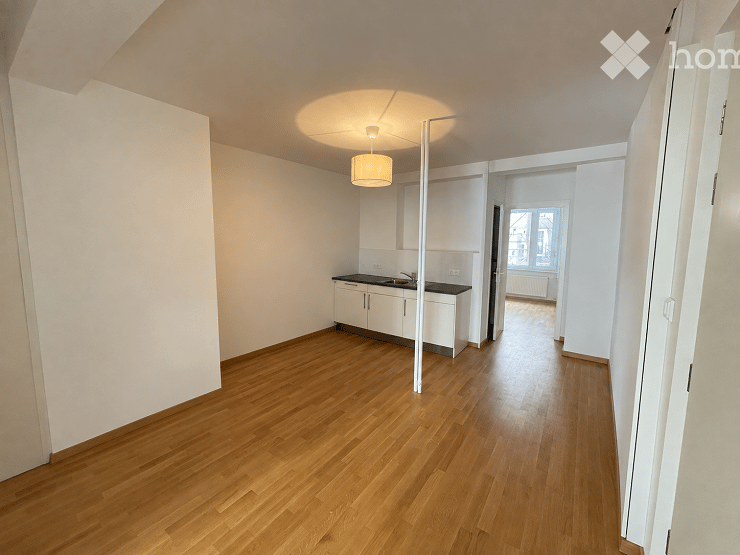 Empty room with light wooden flooring, white walls, and a small kitchen area featuring white cabinets and a sink.