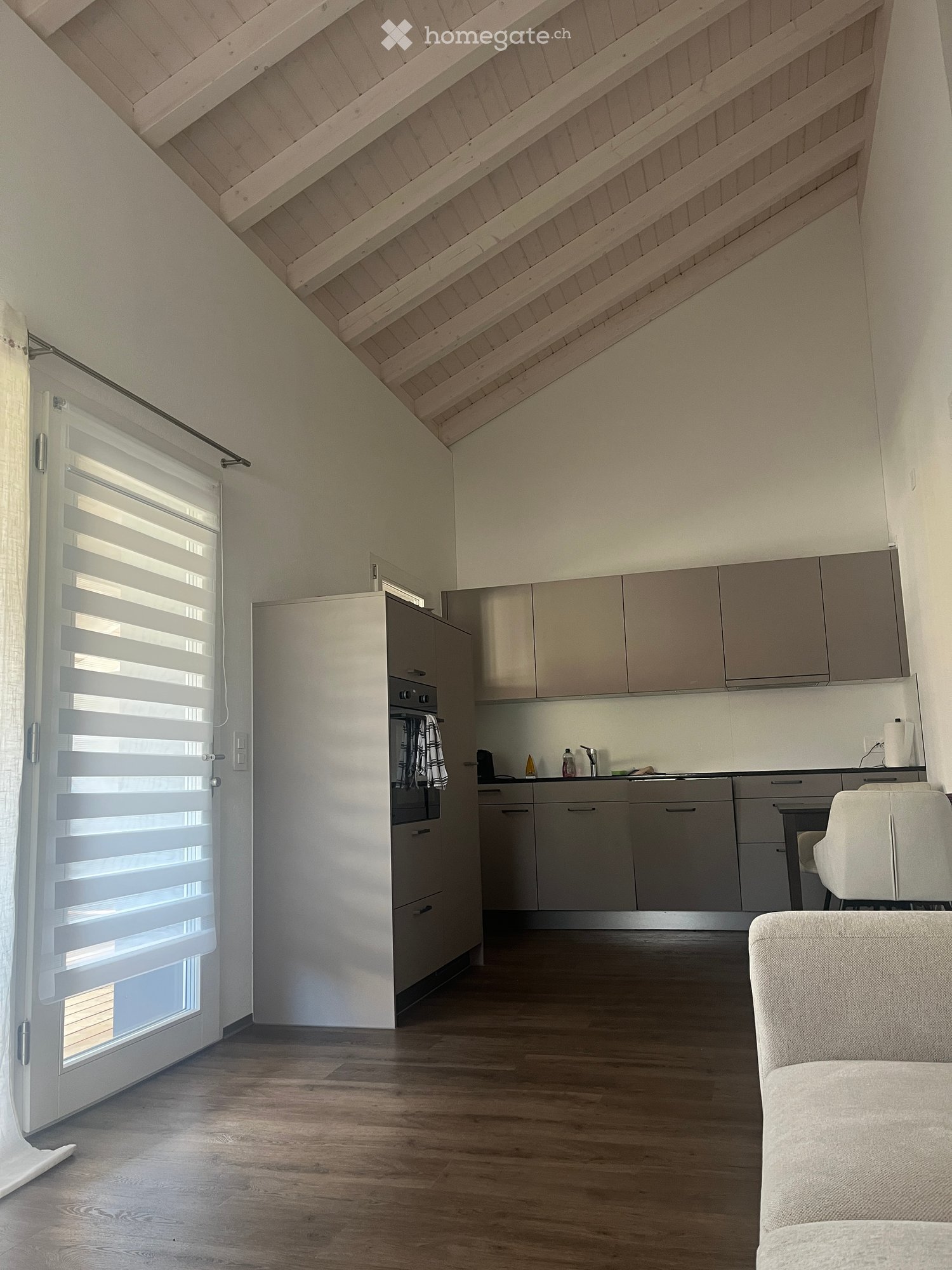 Modern, open-plan living and kitchen area with light wood flooring, grey kitchen cabinets, a white sofa, and a white chair. Features an oven, sink, and blinds on the window.