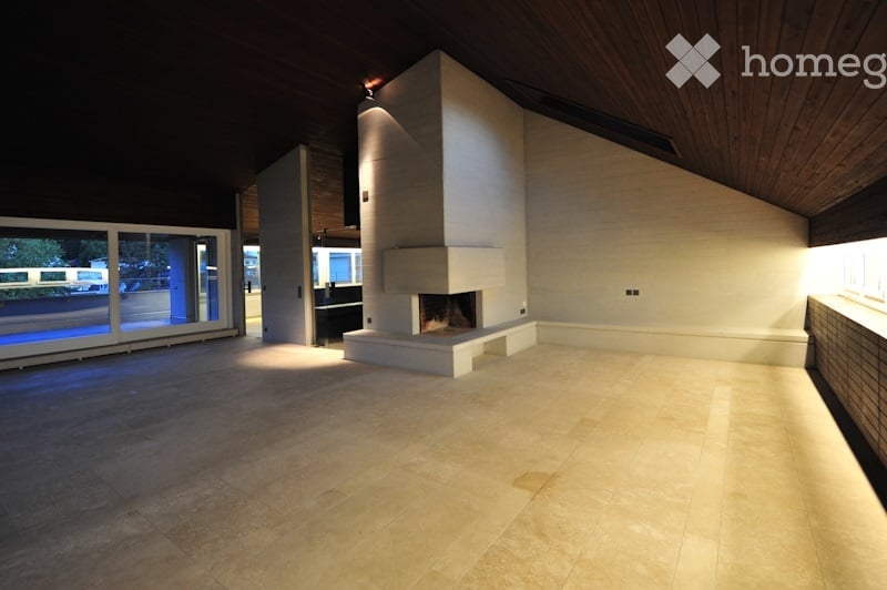 Open-concept space with a raised fireplace, large windows, and wooden ceiling beams. The floor is tiled, and the walls are white.