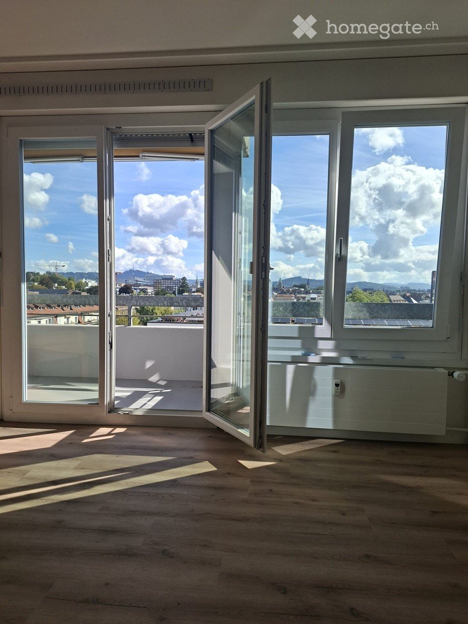 Empty room with wooden flooring, large windows, and open sliding doors leading to a balcony with a cityscape view.