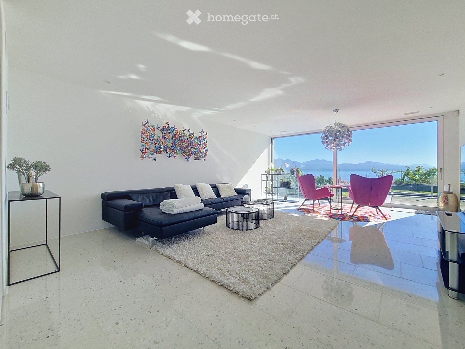 Modern living room with white walls, large windows offering a scenic view, a black sectional sofa, colorful chairs, a coffee table, and a decorative rug.