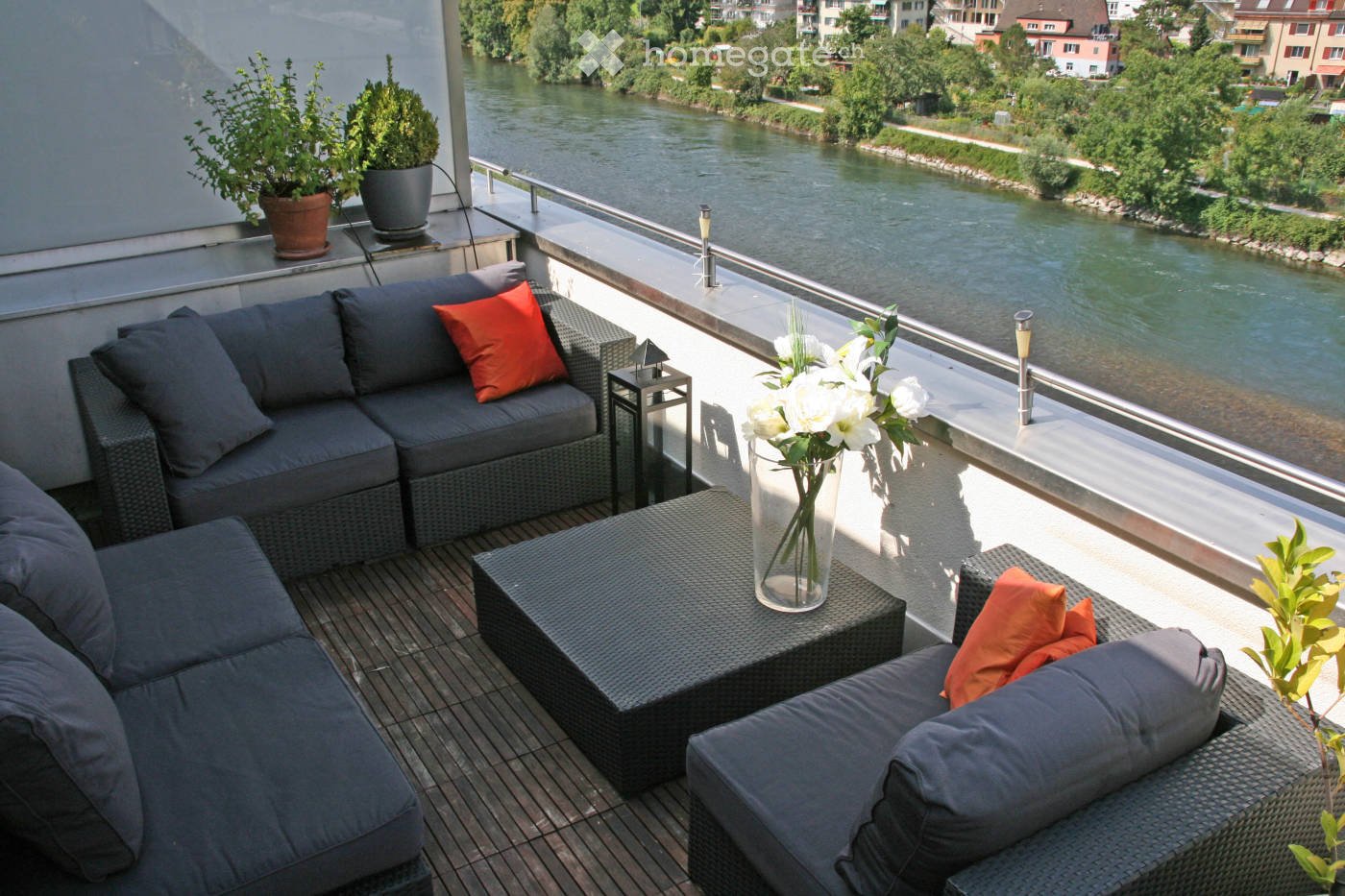 Balcony with a view of a river, featuring a grey wicker sofa, a small table with a vase of white flowers, and potted plants.