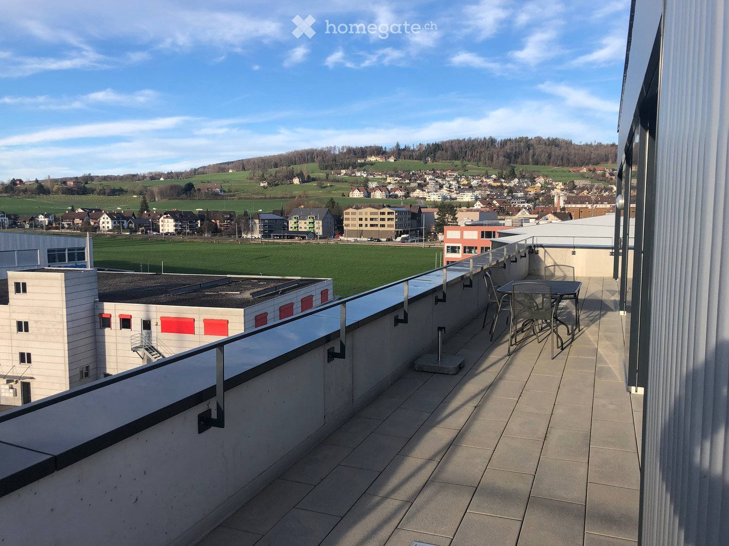 Balcony with tiled floor, metal railing, and a small table with chairs, overlooking a town and green fields.