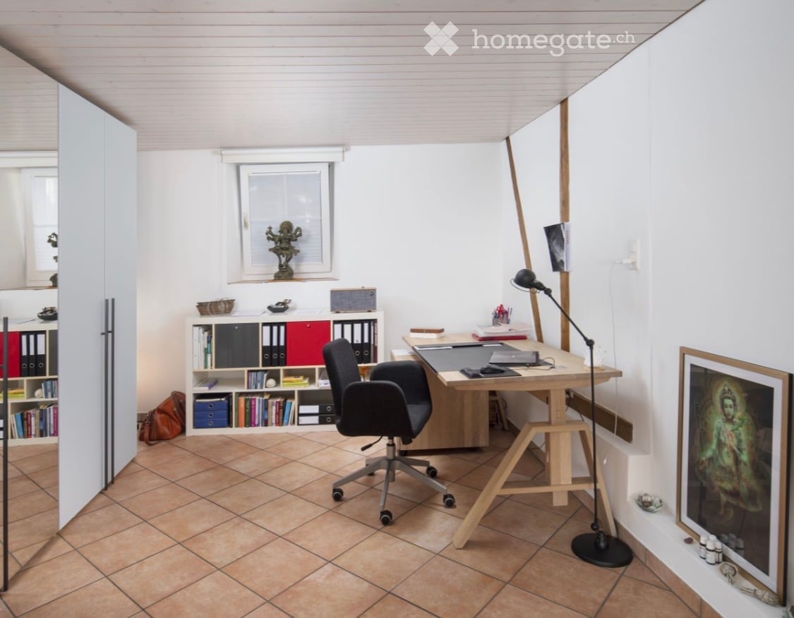 Modern office space with a wooden desk, ergonomic chair, bookshelf, and a large window providing natural light.