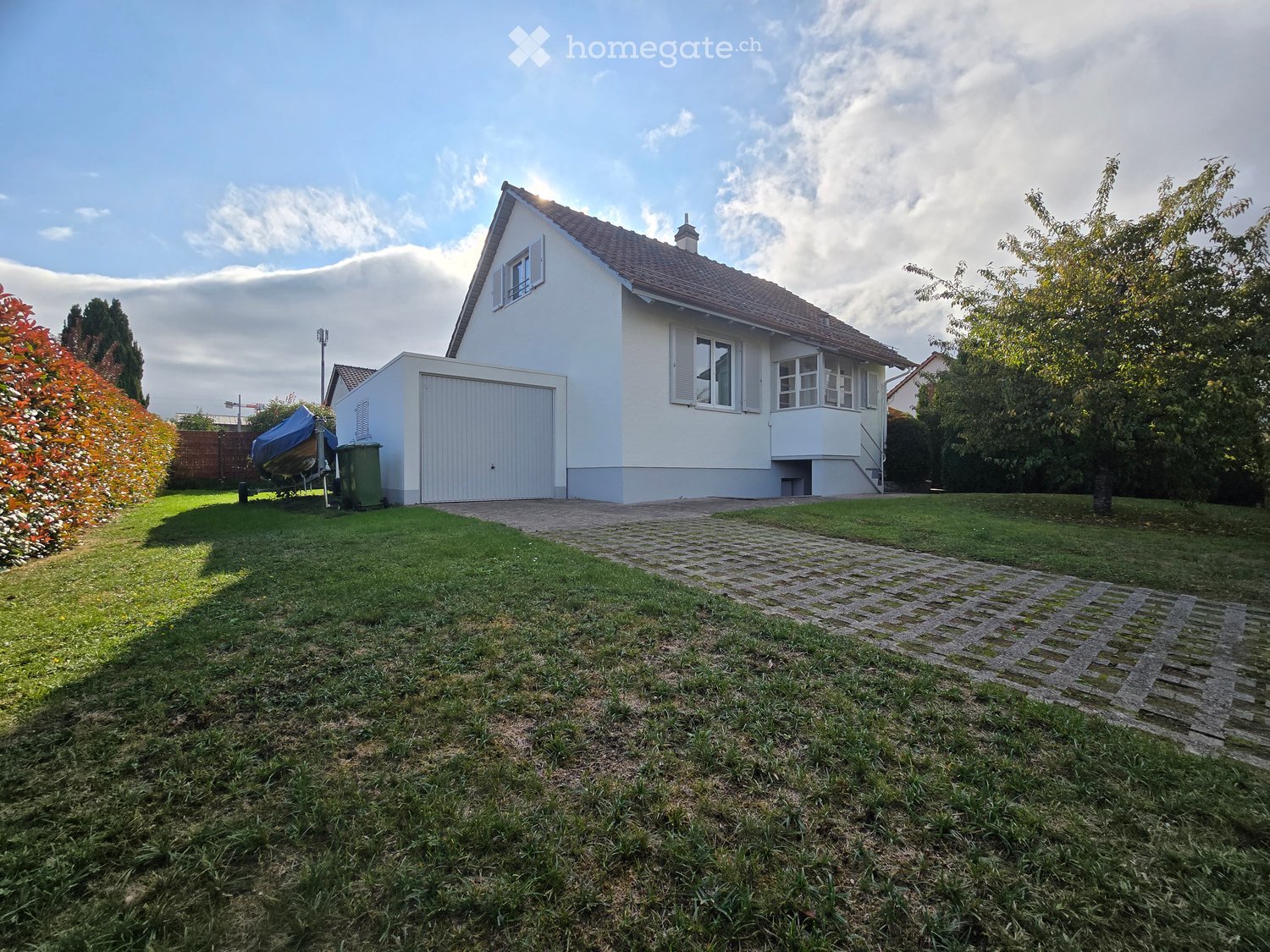 Two-story white house with a garage, surrounded by a well-maintained lawn and garden, with a paved driveway leading to the house.