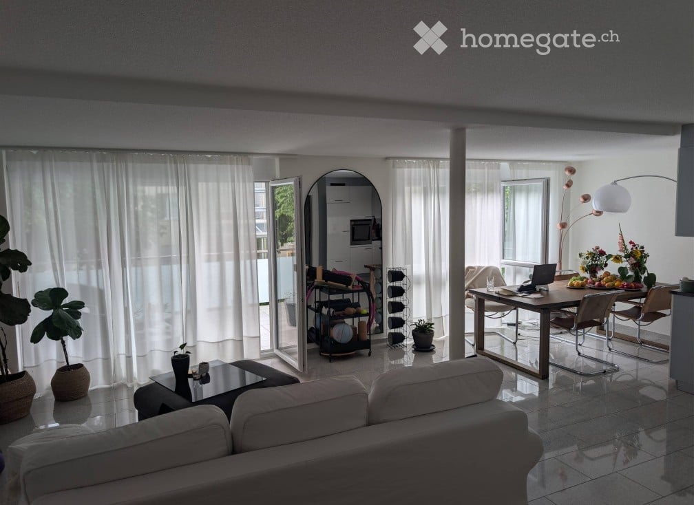 Bright, open-plan living and dining area with large windows and sliding glass doors leading to a balcony. Features a white sofa, coffee table, dining table with chairs, laptop, flower vase, floor lamp, wine rack, and mirror.