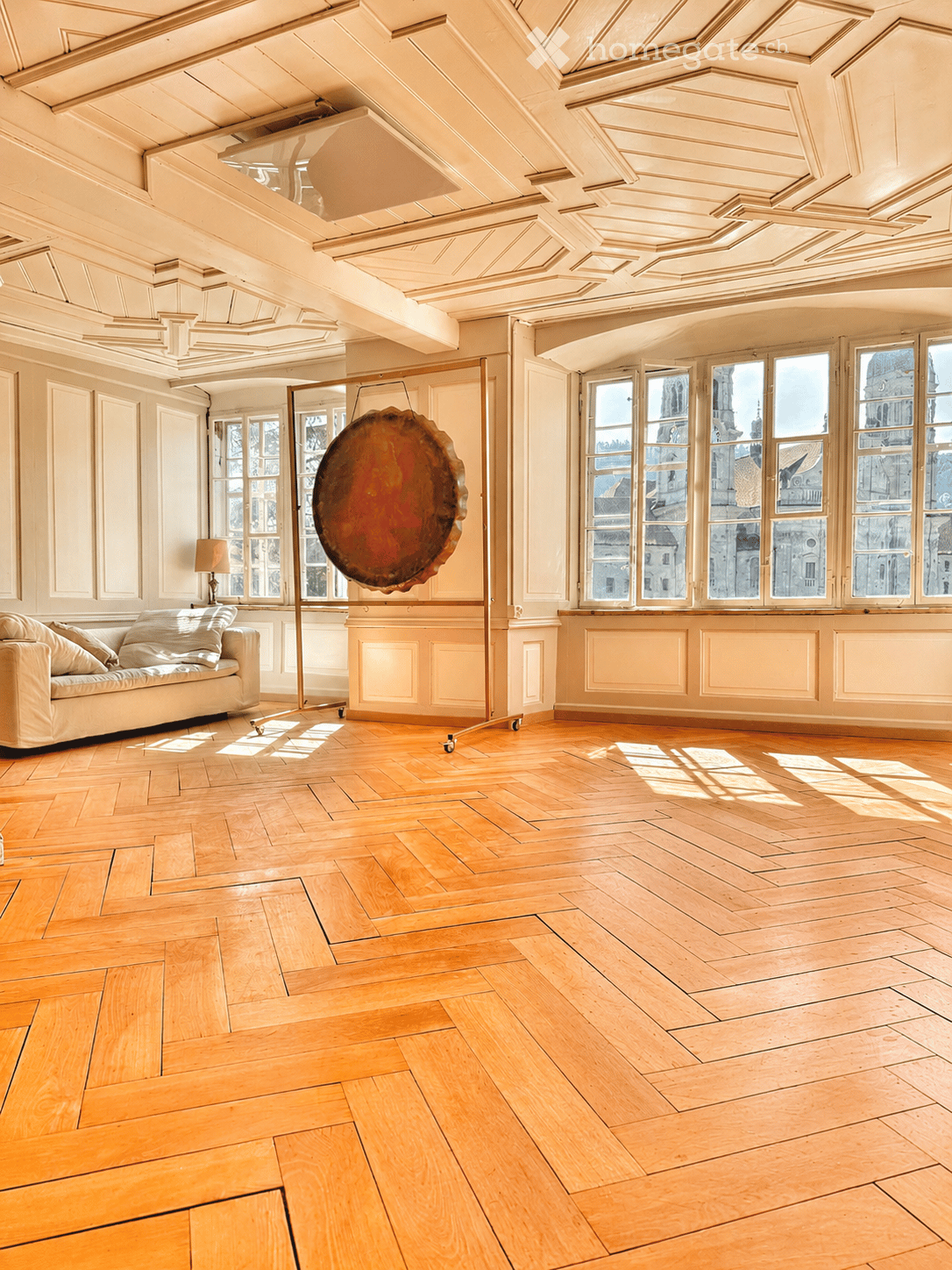 Bright, spacious room with wooden herringbone flooring, large windows offering a city view, a cream-colored sofa, a table lamp, and a large decorative gong suspended from the ceiling.