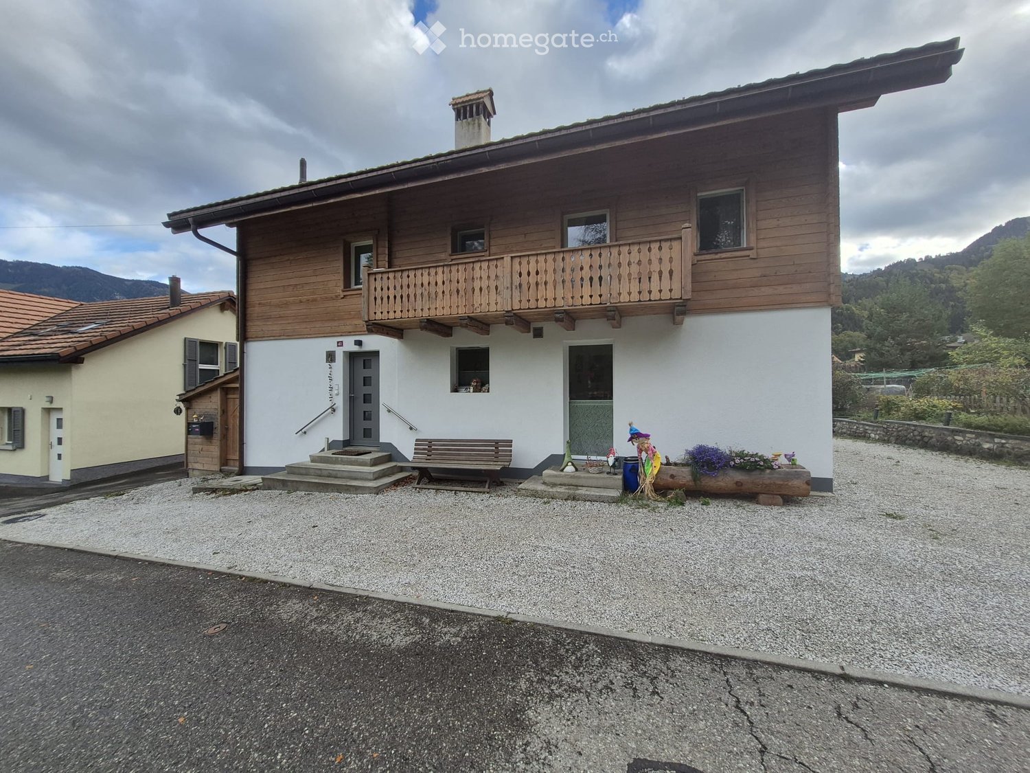 Two-story wooden house with a balcony, surrounded by a gravel driveway and small garden decorations.