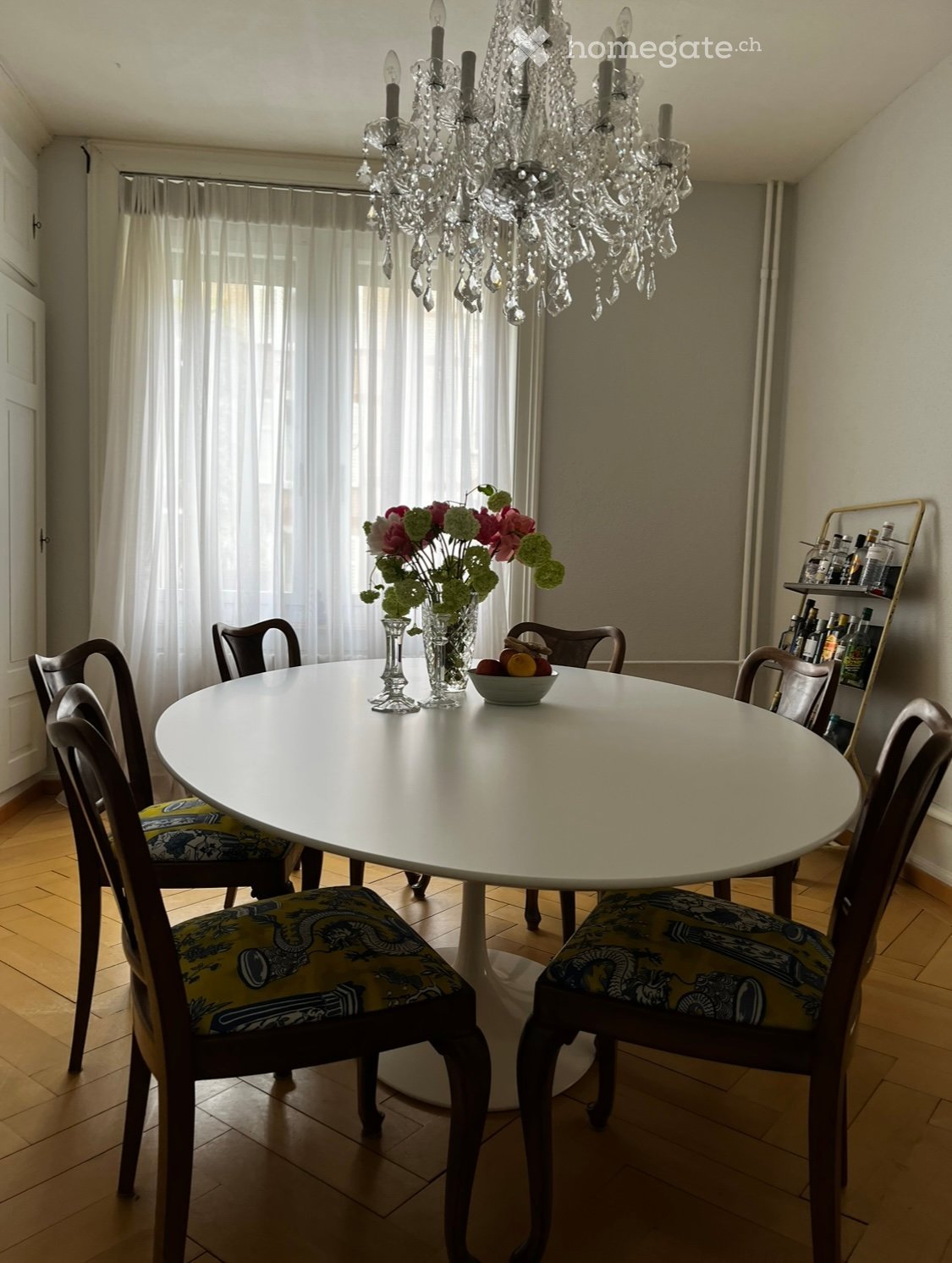 Dining room with a round white table surrounded by six chairs with patterned cushions, a chandelier, a vase with flowers, and a shelf with bottles.
